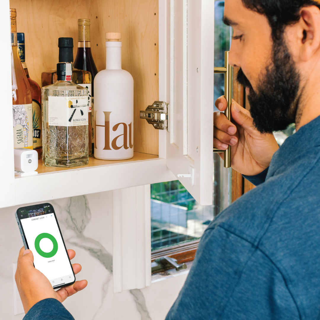 Ein Mann öffnet einen Schrank mit Alkoholflaschen. Er hält ein Telefon, das ein entsperrtes Schrankschloss anzeigt. Der Schrank hat eine weiße Tür.