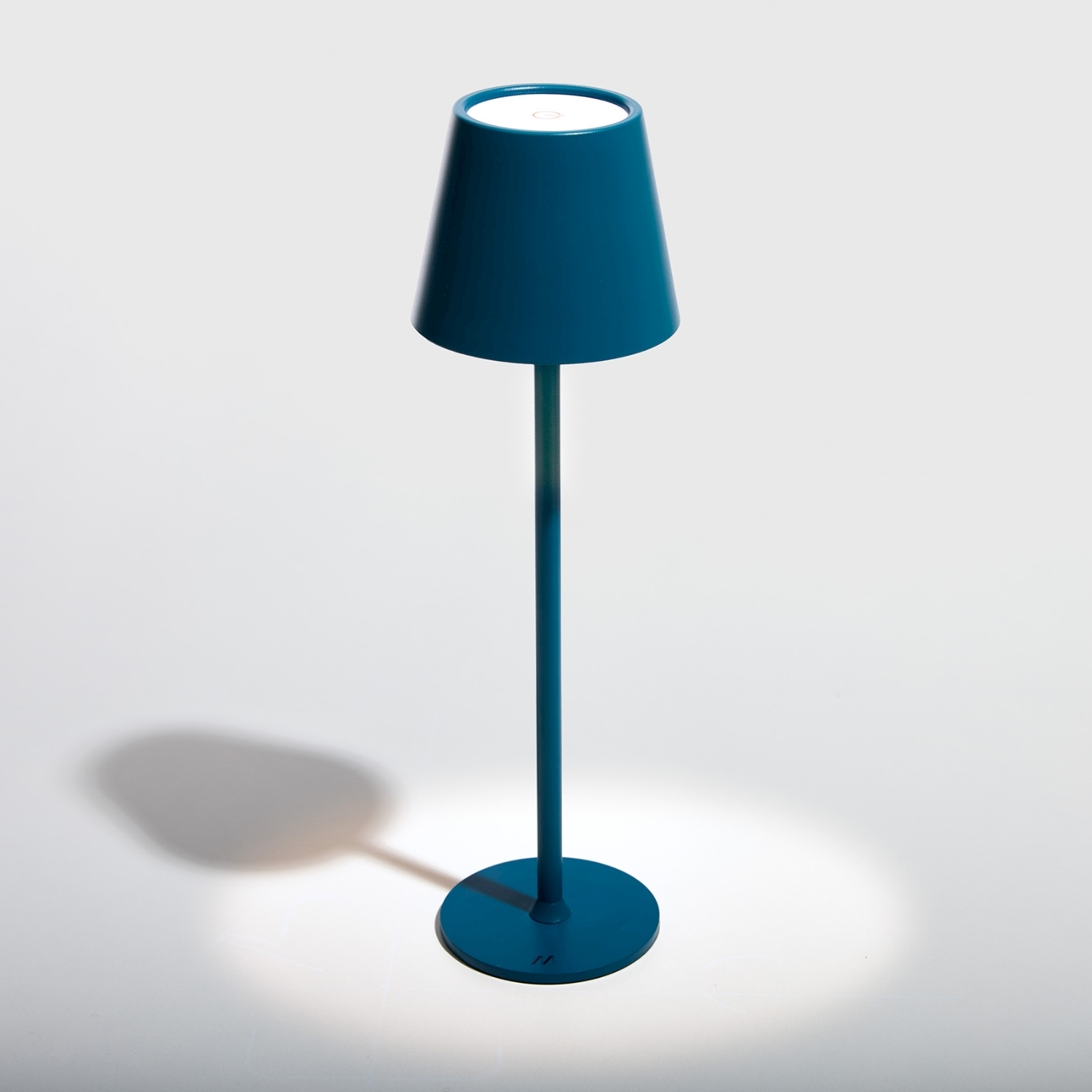 Una lampada turchese è su una superficie bianca. La lampada ha una base rotonda, un lungo stelo e un paralume. Si proietta un'ombra.