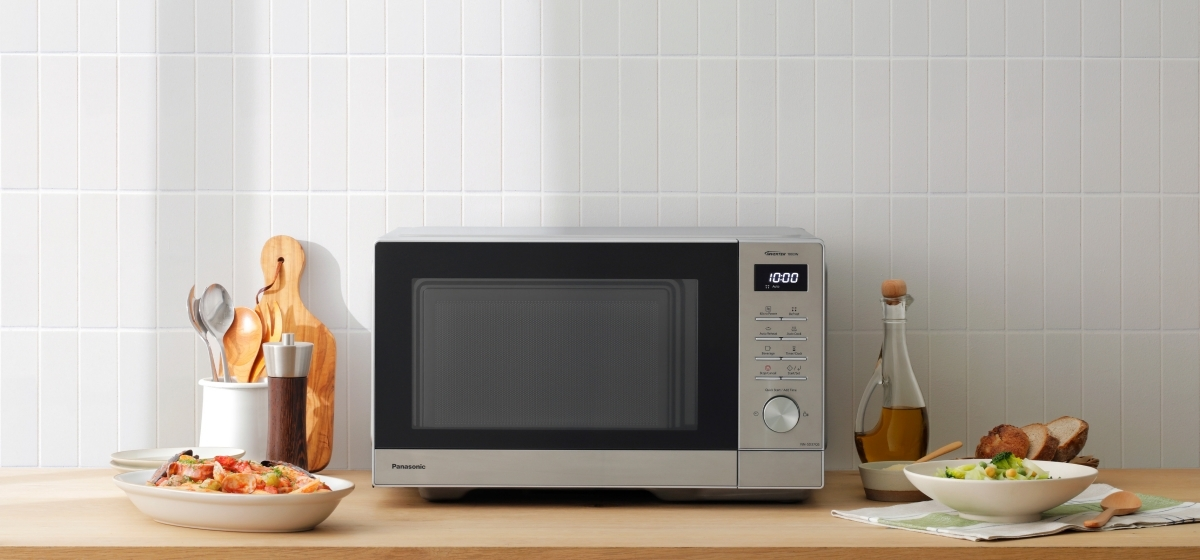 Kuchenka mikrofalowa stoi na drewnianej powierzchni. Otaczają ją jedzenie i przybory kuchenne. Biała ściana z płytek w tle.