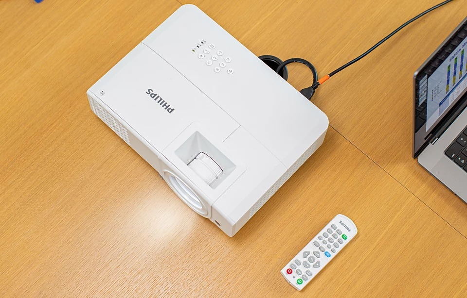 Biały projektor z pilotem i laptopem na drewnianym stole. Projektor jest podłączony do laptopa.