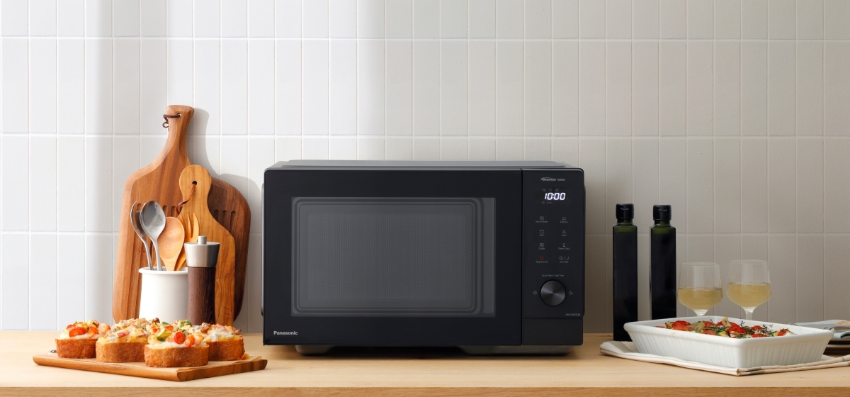 Czarna kuchenka mikrofalowa stoi na drewnianym blacie. Tosty, oliwa z oliwek i kieliszki do wina na blacie. Białe kafelki w tle.