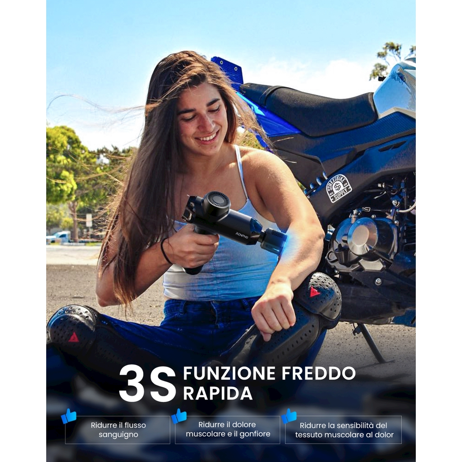 Una donna usa una pistola massaggiante sul braccio vicino a una moto blu. Indossa jeans e ginocchiere.
