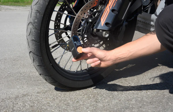 Eine Person, die den Reifendruck eines Motorrads mit einem digitalen Messgerät überprüft. Reifen, Rad und Gabel sind sichtbar. Das Messgerät zeigt 30,0.