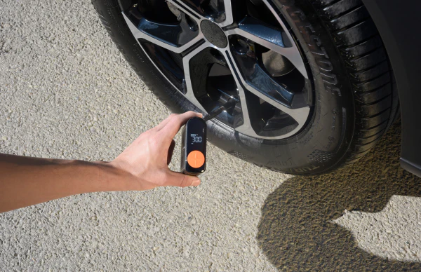 Eine Hand hält ein Reifendruckmessgerät neben einem Autoreifen. Das Messgerät zeigt 300 an. Das Auto steht auf einem Bürgersteig.
