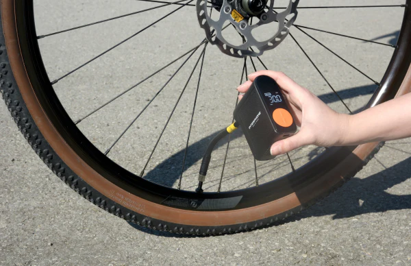 Eine Person pumpt einen Fahrradreifen mit einer schwarz-orangen Pumpe auf. Der Reifen ist braun und das Rad ist schwarz. Der Hintergrund ist grau.