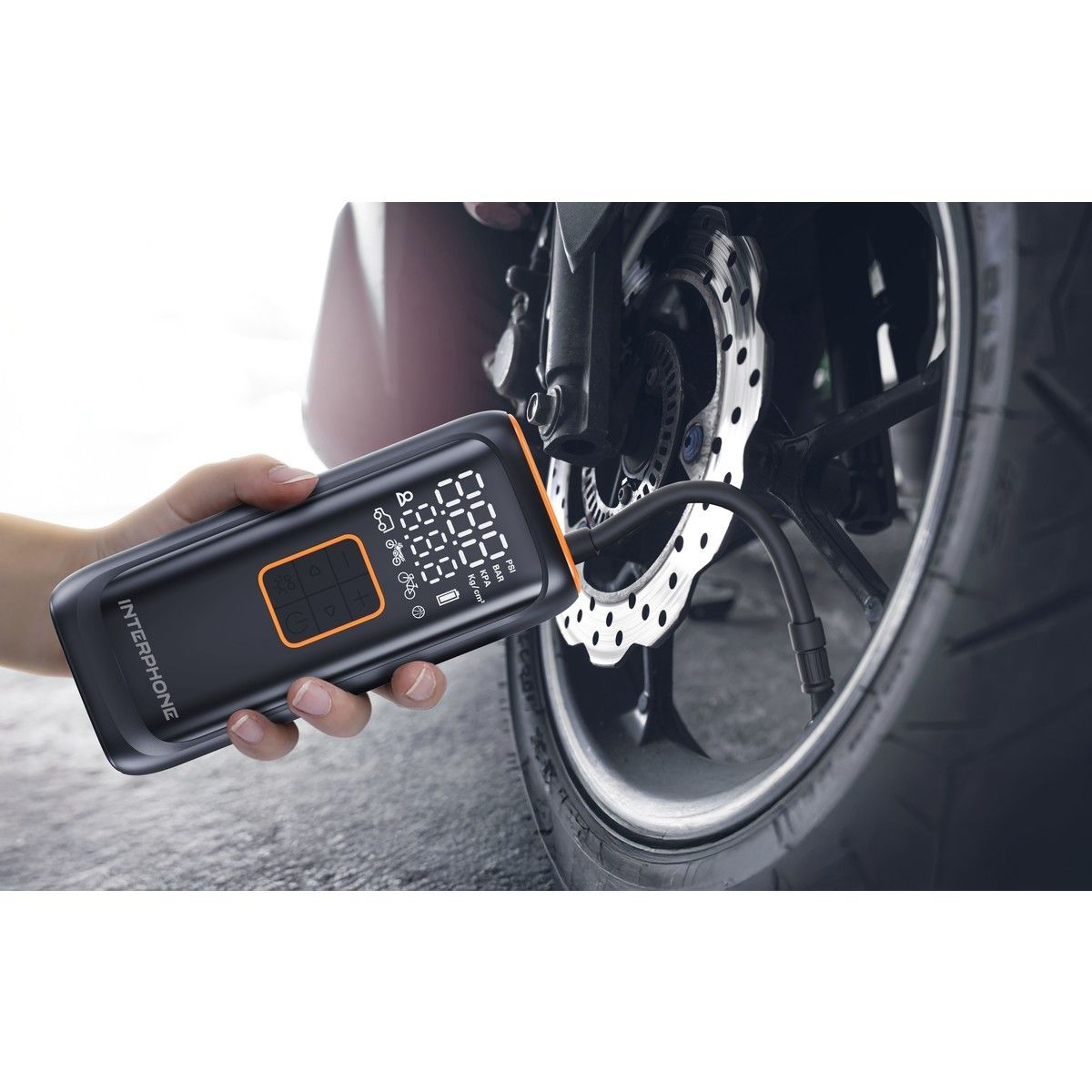 Eine Hand hält eine schwarz-orangefarbene Interphone-Luftpumpe, die mit einem Motorradreifen verbunden ist. Das digitale Display zeigt Druckwerte an.