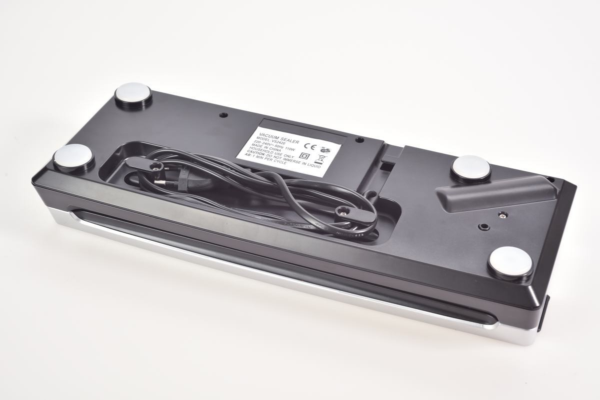 Czarna zgrzewarka próżniowa, ze srebrnym wykończeniem, czarnym przewodem i białymi nóżkami, na białej powierzchni. Widoczna etykieta.