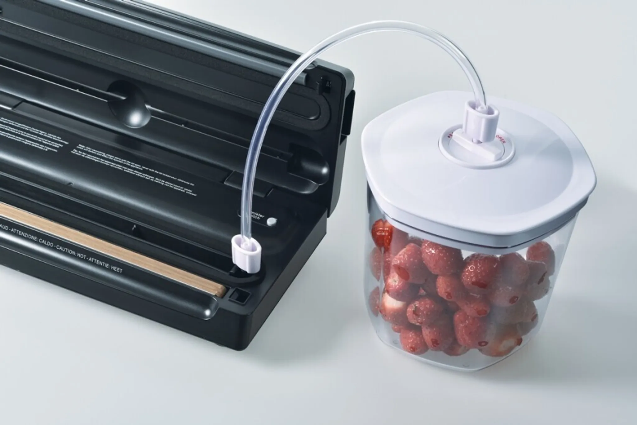 Zgrzewarka próżniowa z pojemnikiem truskawek połączonym przezroczystą rurką. Zgrzewarka jest czarna, a pojemnik przezroczysty z białą pokrywką.