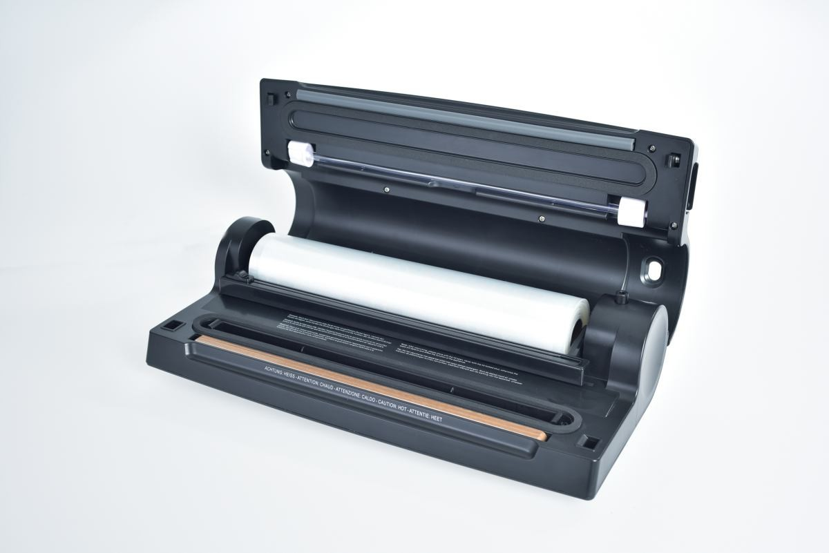 Czarna zgrzewarka próżniowa z rolką przezroczystego plastiku w środku, na białej powierzchni. Otwarta góra, z napisem 'Uwaga: Gorąco'.