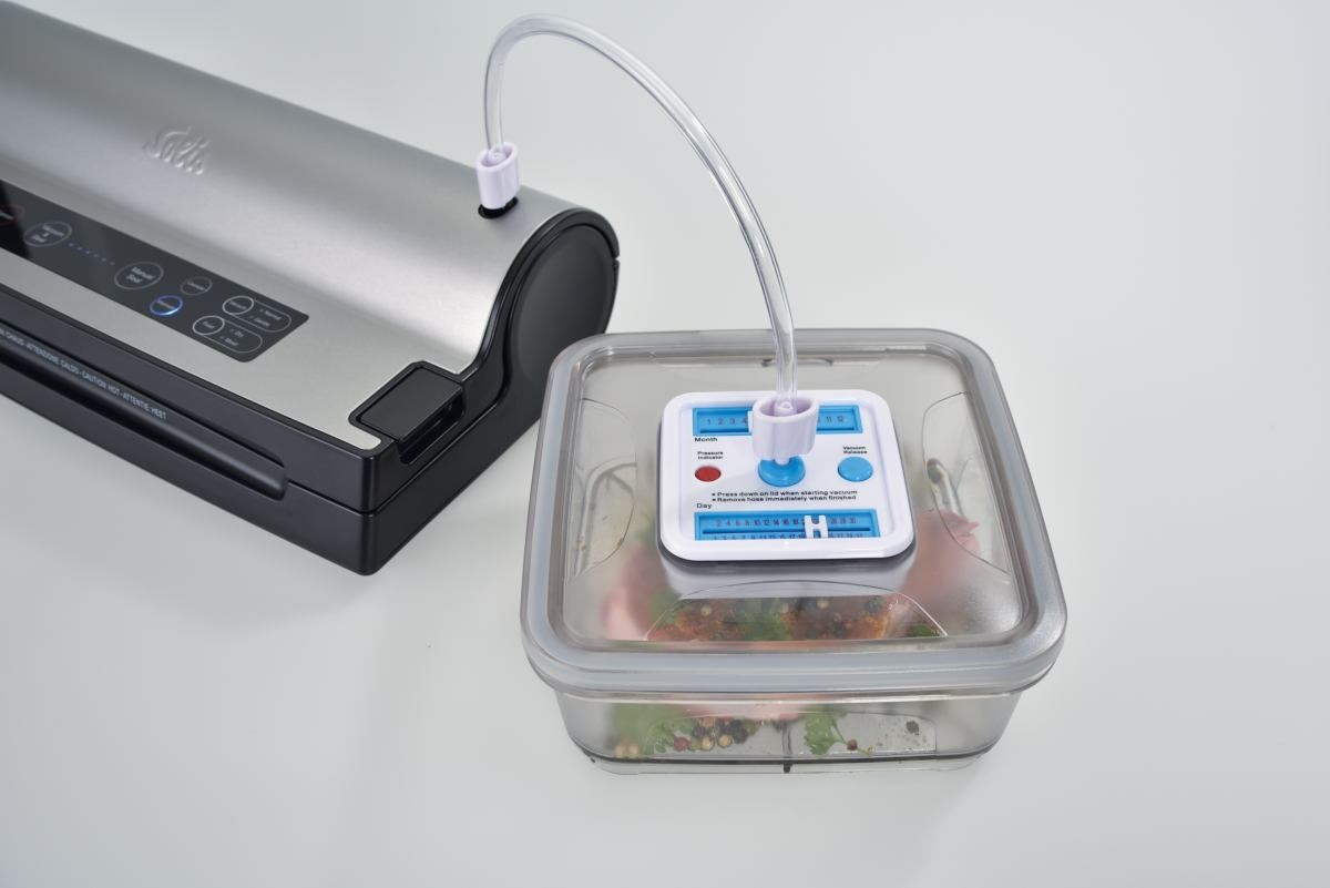 Zgrzewarka próżniowa połączona z pojemnikiem z jedzeniem. Przezroczysta rurka łączy zgrzewarkę i pojemnik. Pojemnik jest kwadratowy.