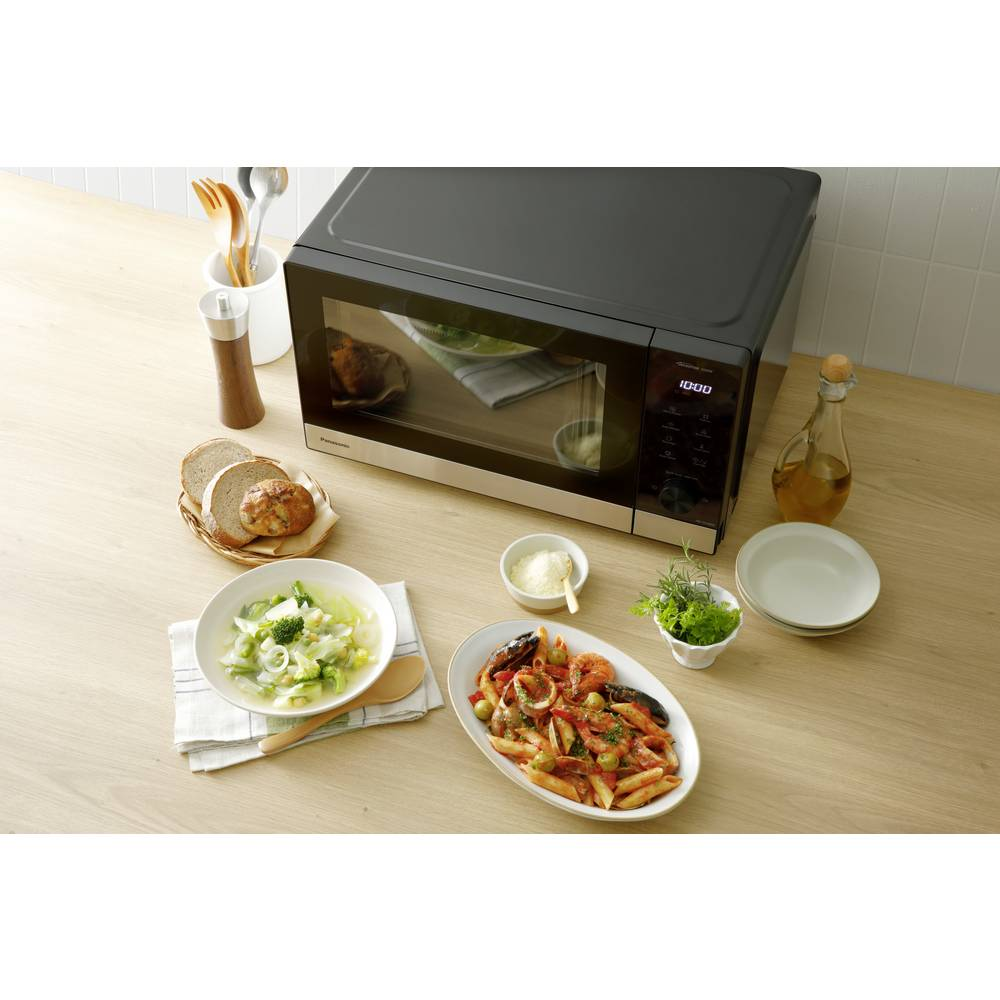 Kuchenka mikrofalowa stoi na drewnianej powierzchni z jedzeniem, takim jak zupa, makaron, chleb i olej, sugerując kuchnię.