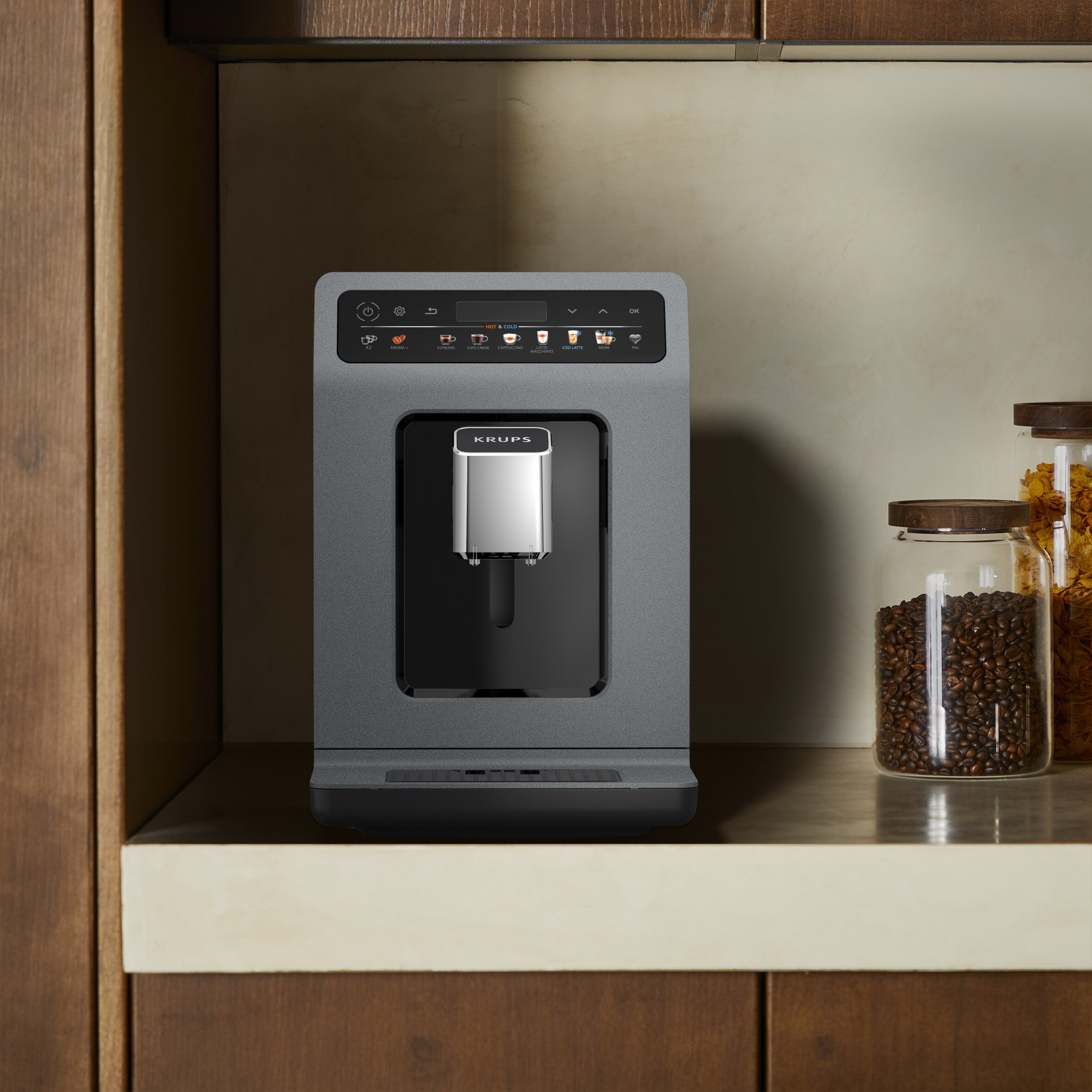 Ekspres do kawy Krups na półce, z dwoma słoikami ziaren kawy i płatków zbożowych. Maszyna ma wyświetlacz.