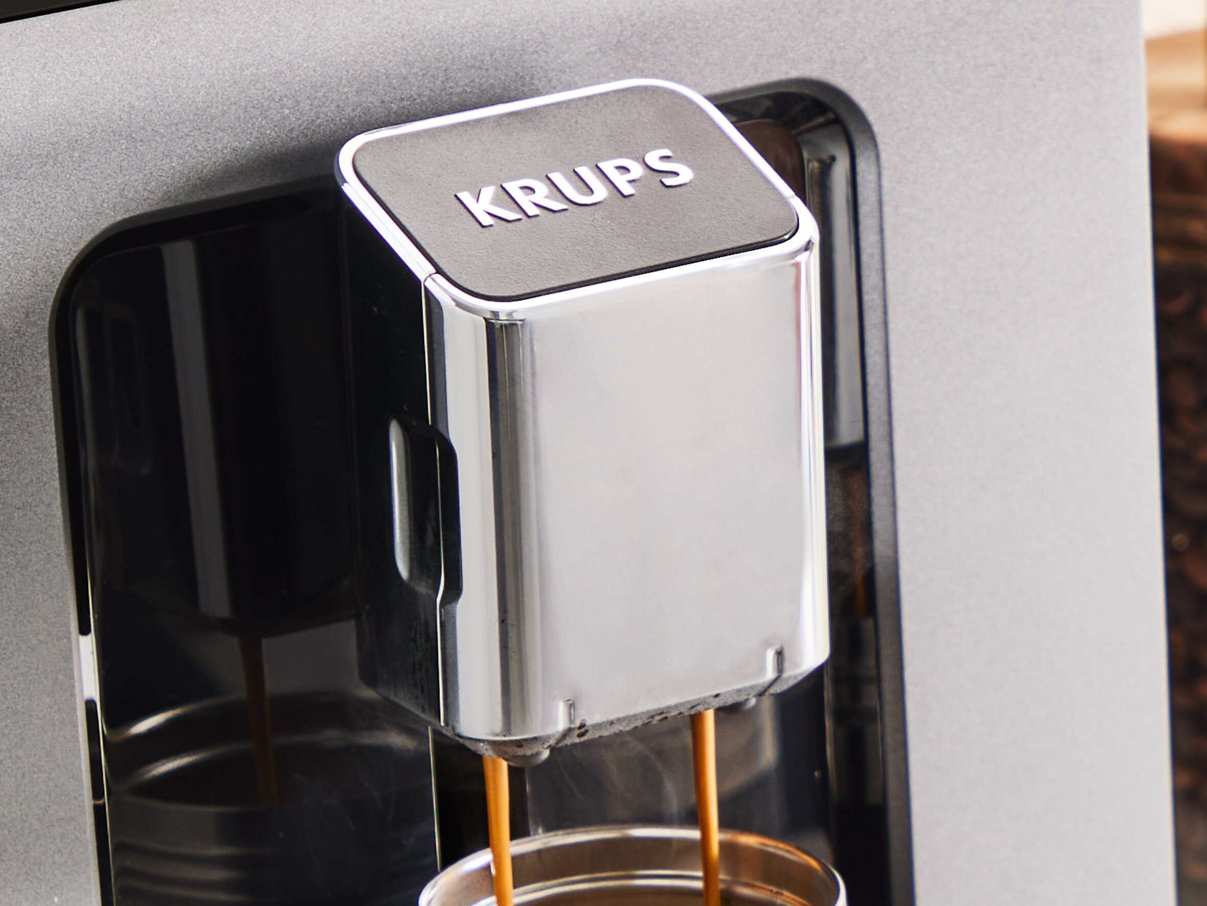 Ekspres do kawy Krups. Kawa wlewana jest do filiżanki. Maszyna jest srebrna i czarna.