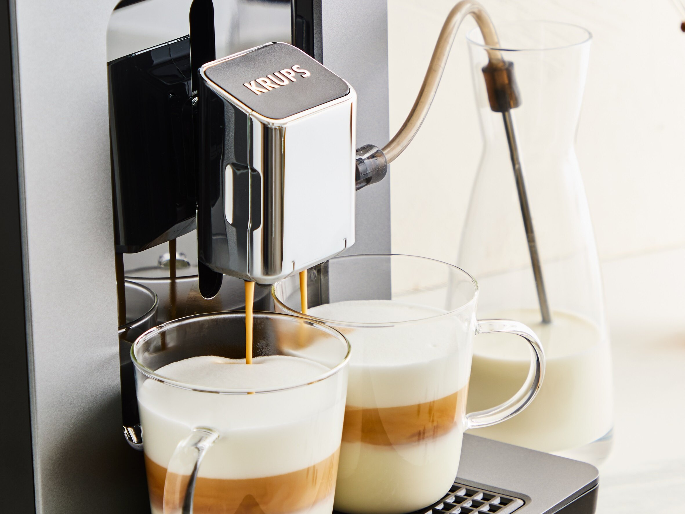 Ekspres do kawy dozuje kawę do szklanych kubków z warstwowym mlekiem. W tle pojemnik na mleko.