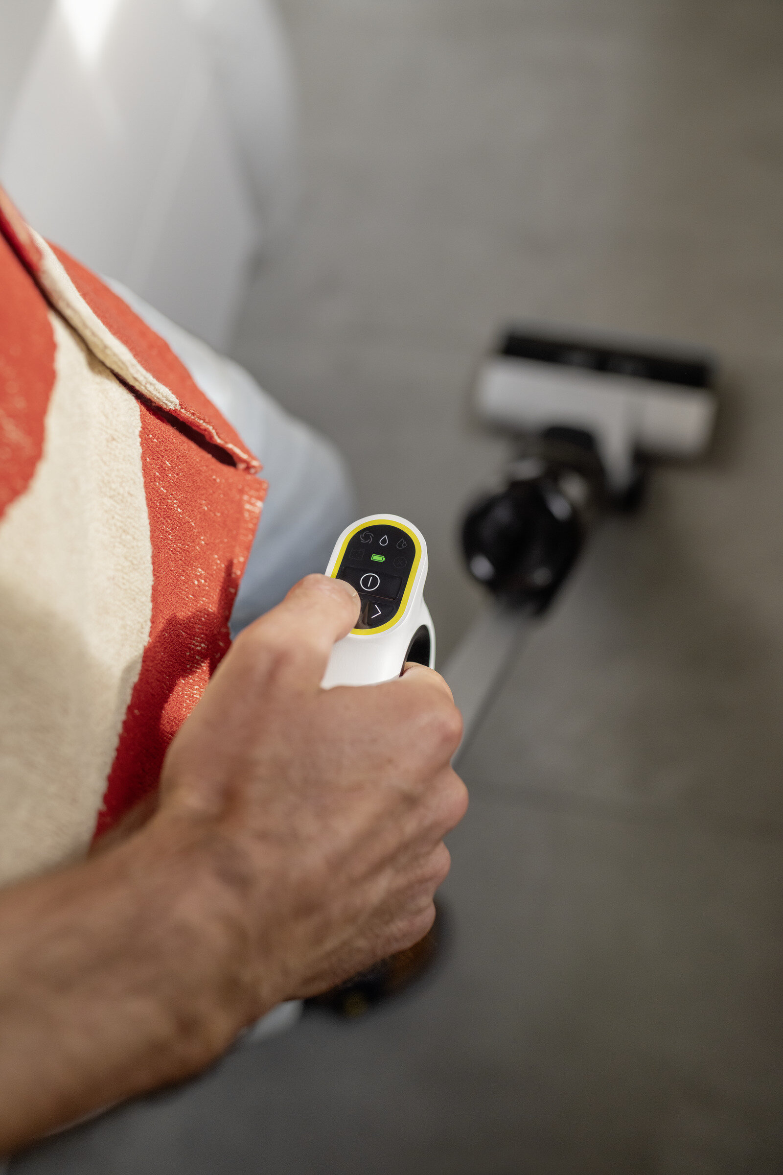 Ręka osoby trzymająca biały kontroler odkurzacza. Tło przedstawia odkurzacz i powierzchnię we wzory.