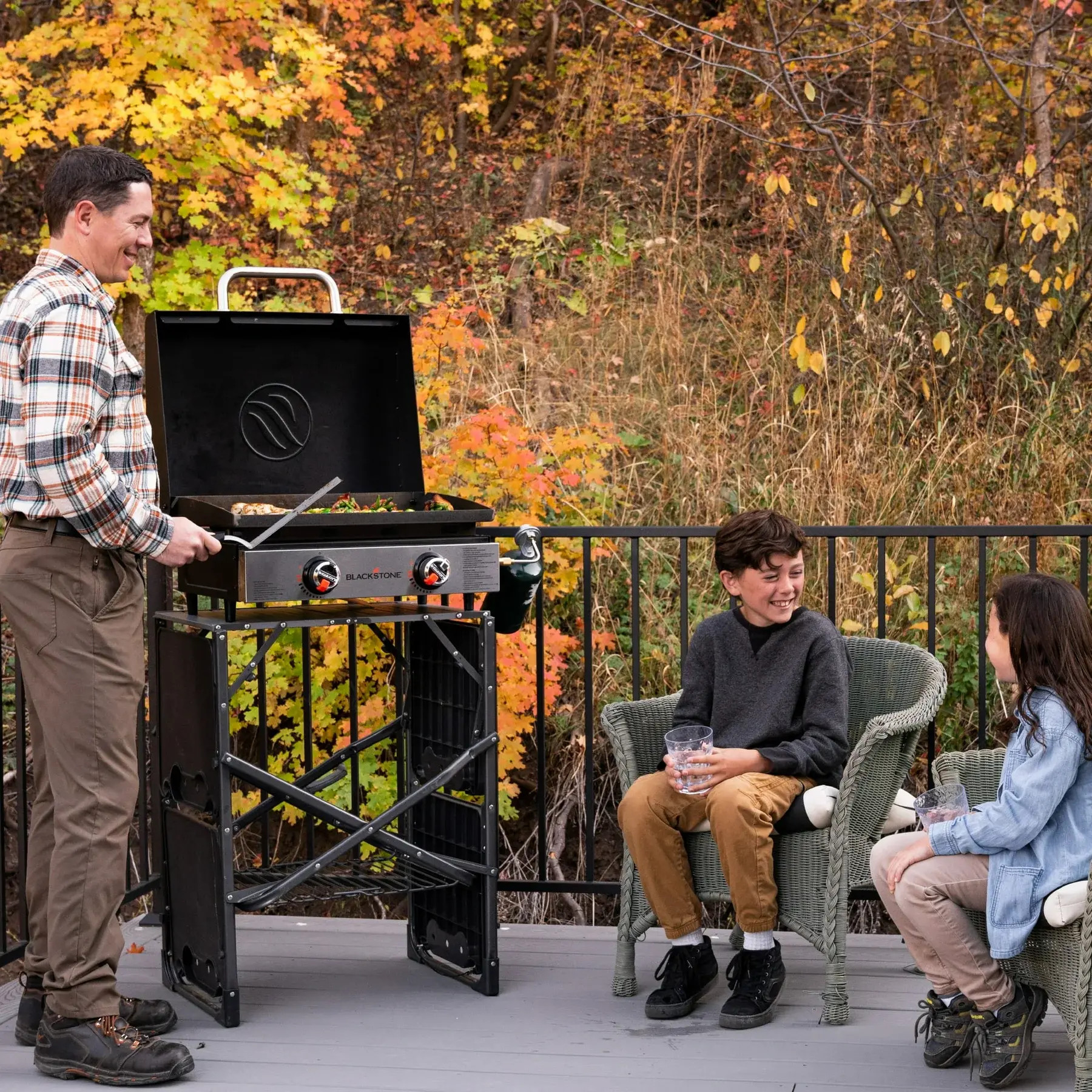 Mężczyzna grilluje na zewnątrz, a dwoje dzieci patrzy. Jesienne liście w tle. Grill jest czarny.