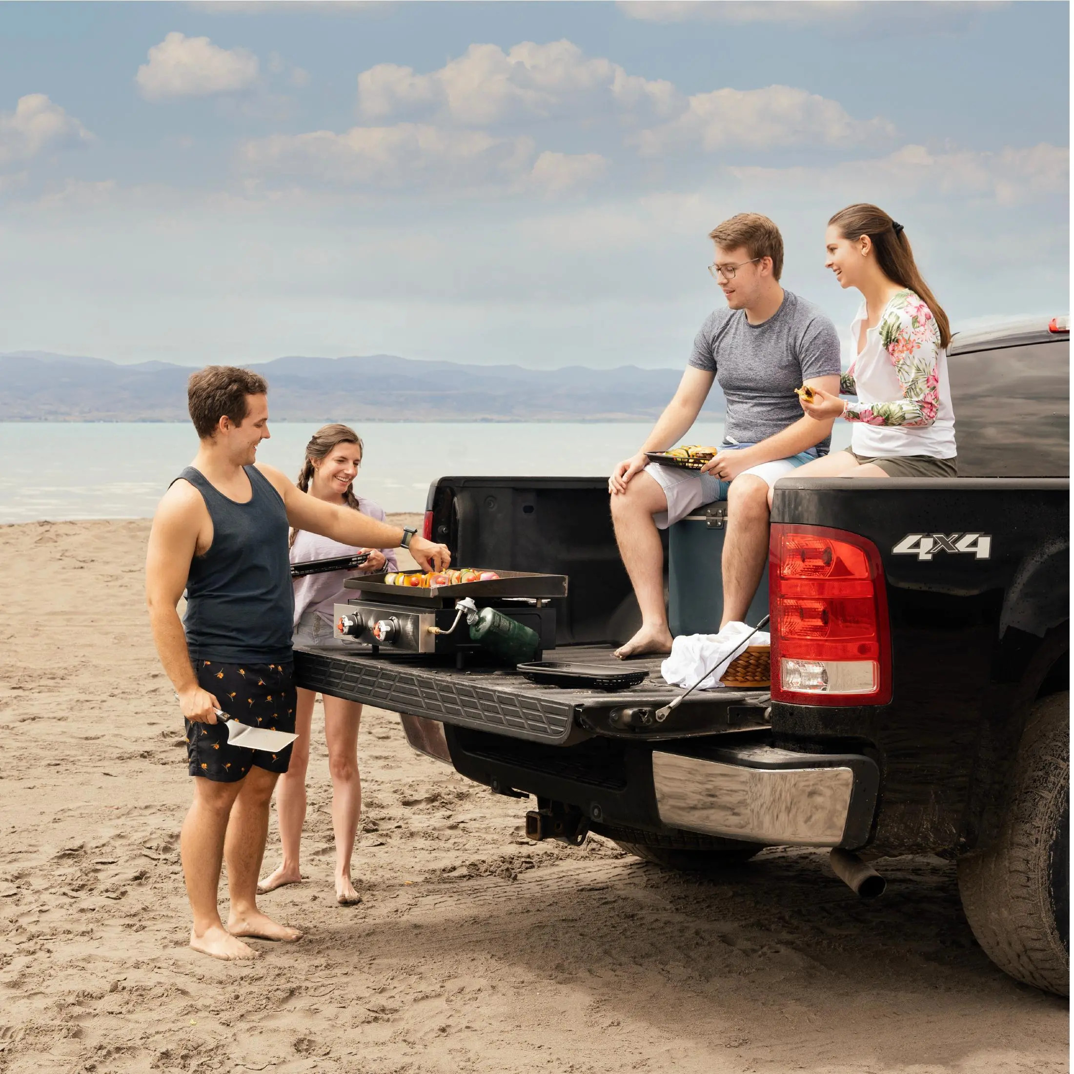 Ludzie grillują na pace ciężarówki na plaży. Dwóch siedzi na pace, dwóch grilluje, a jeden trzyma szpatułkę.