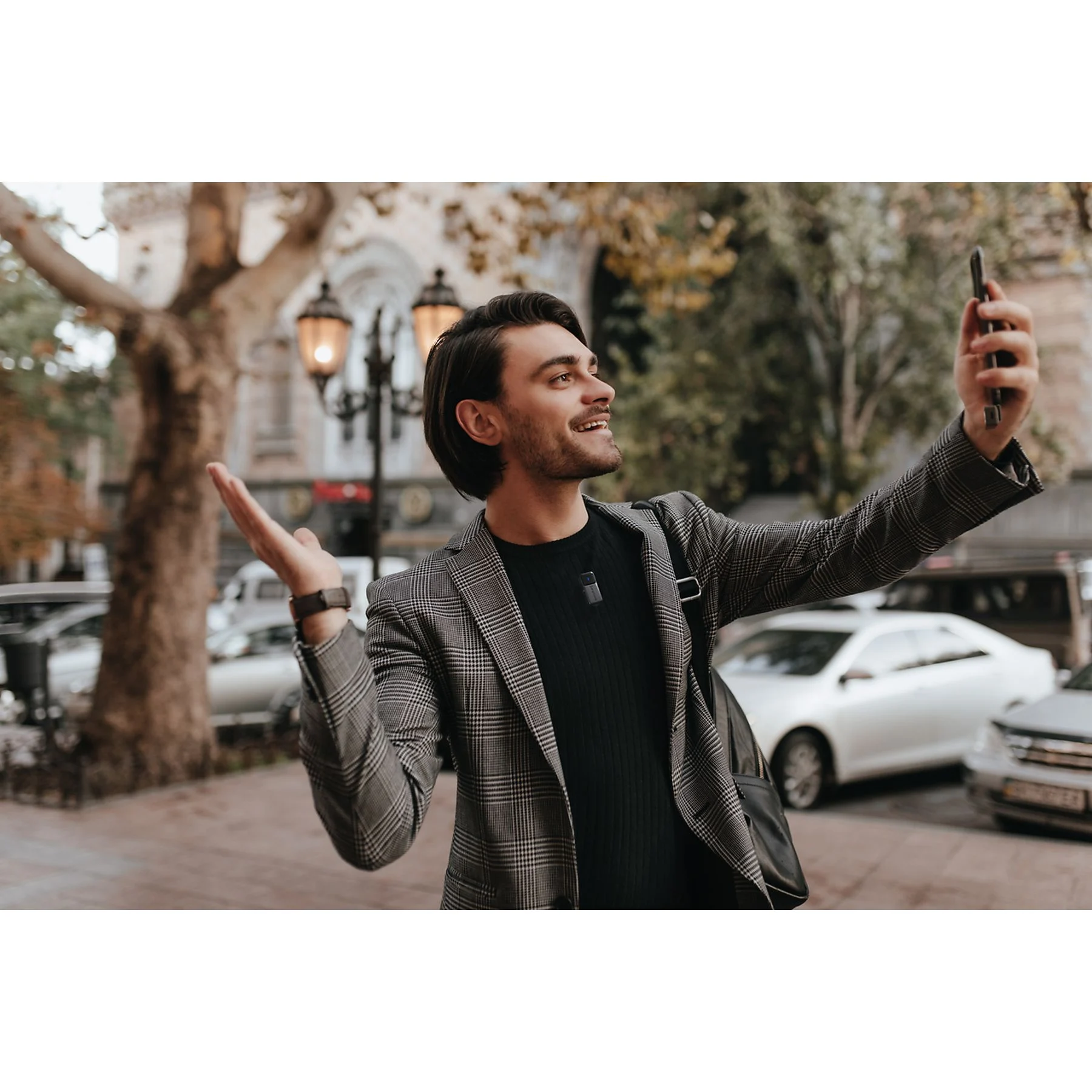 Ekose ceketli bir adam telefonla selfie çekiyor, gülümsüyor. Kaldırımda dışarıda.