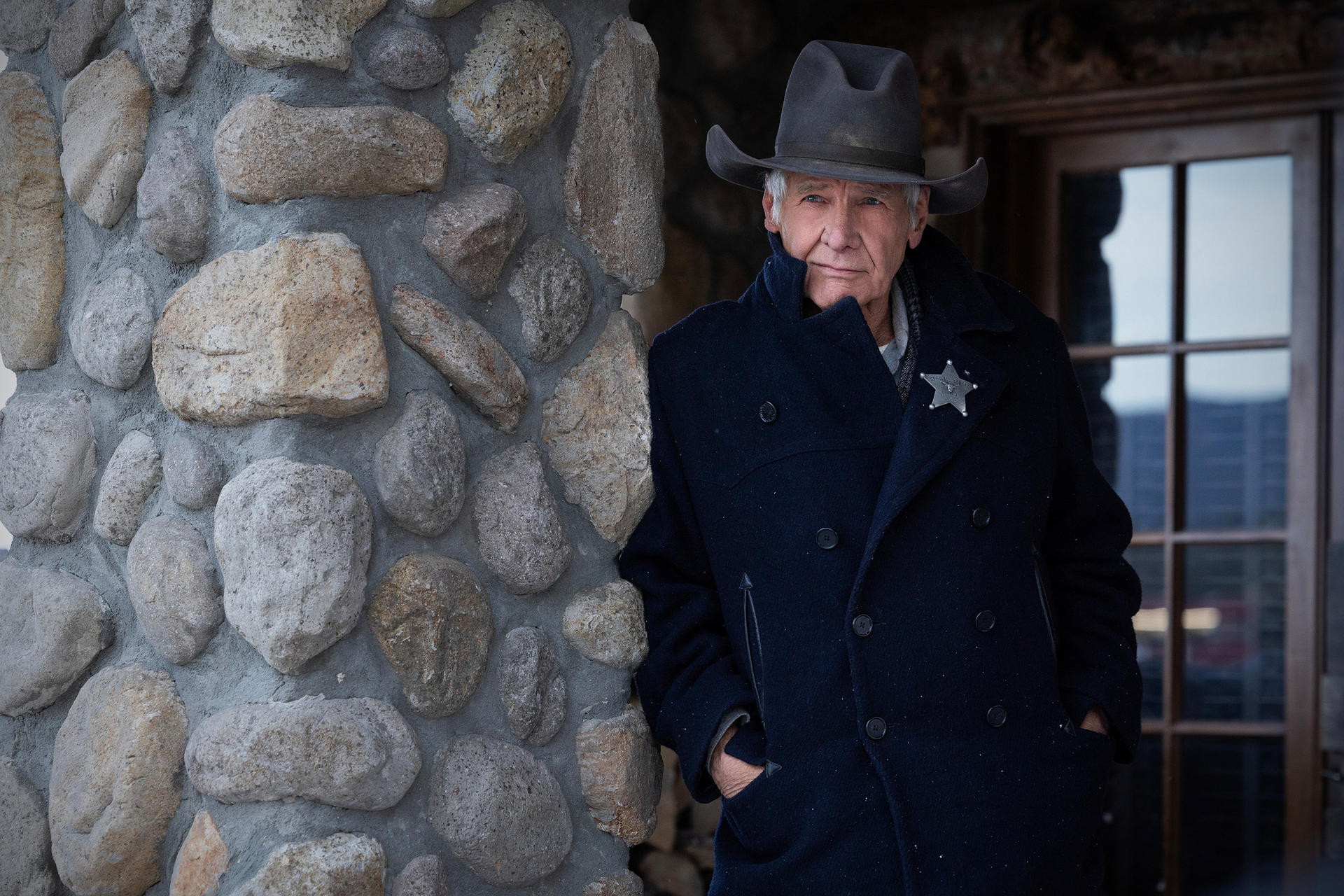 Harrison Ford in Cowboyhut und Mantel lehnt an einer Steinmauer. Ein Sheriff-Abzeichen ist an seinem Mantel befestigt.