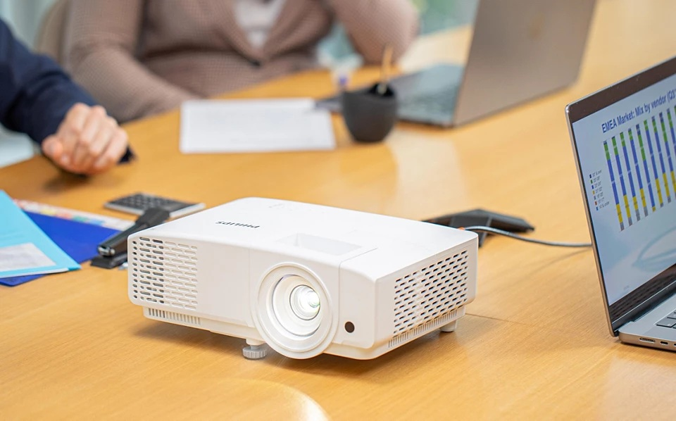 Biały projektor na drewnianym stole. Laptop wyświetla wykres. Ludzie siedzą wokół stołu. Spotkanie.