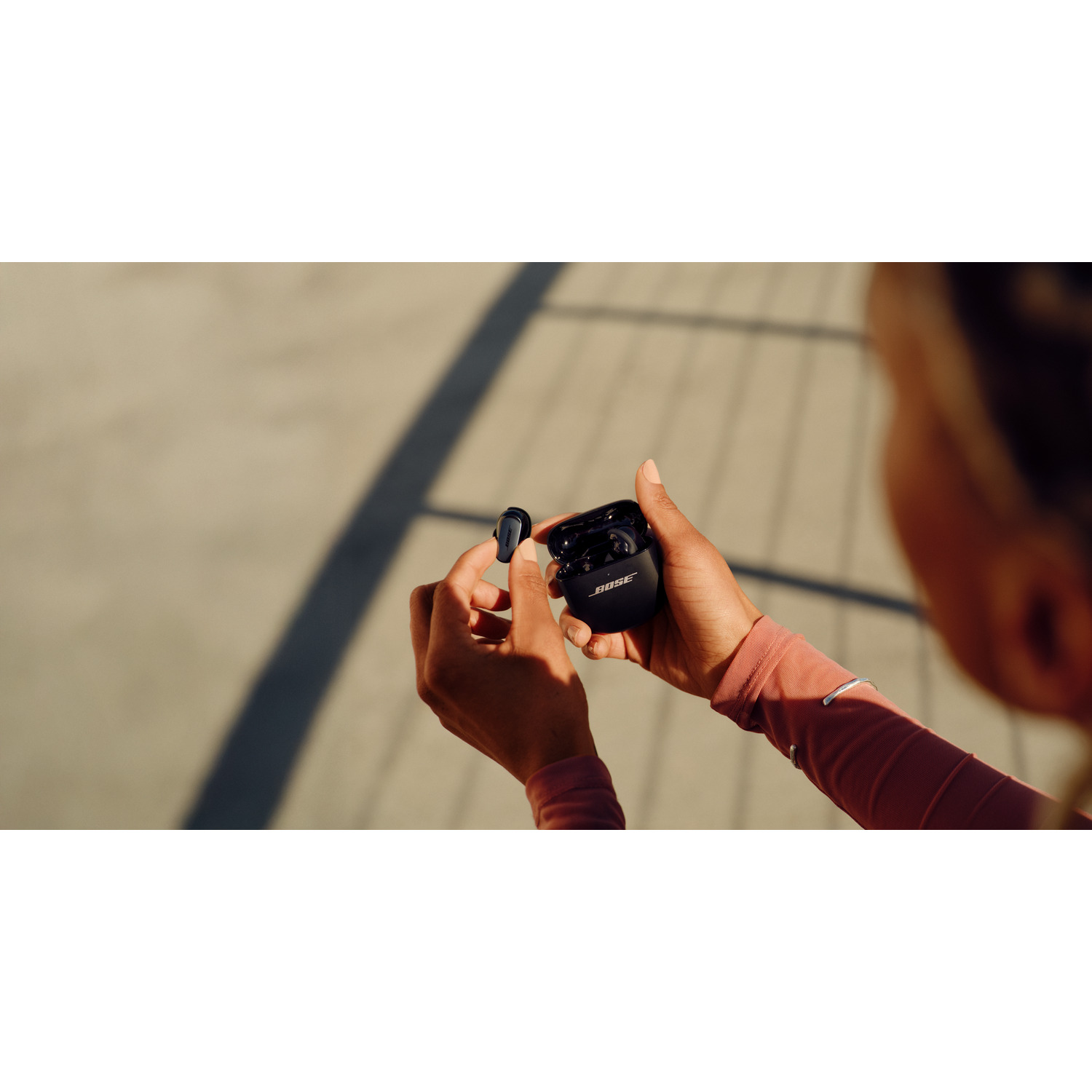 A person holds black Bose earbuds and their charging case. The background is a blurred outdoor scene with shadows.