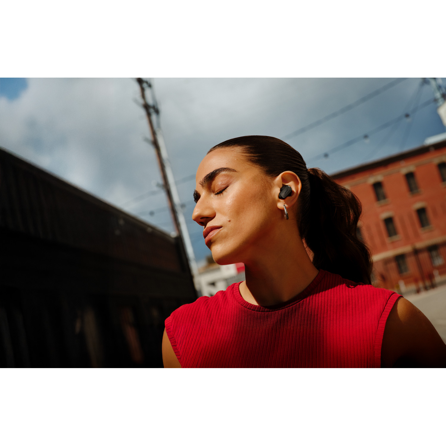 A woman with closed eyes wears earbuds. She is in a red top. Background shows a building and a cloudy sky.