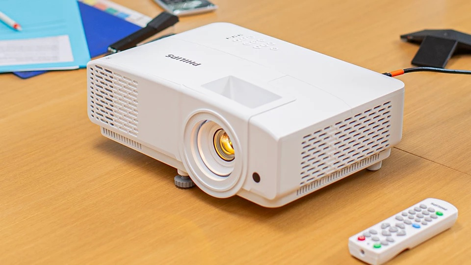 Biały projektor Philips na drewnianym stole. Pilot zdalnego sterowania jest na pierwszym planie. Papiery i czarny telefon są w tle.