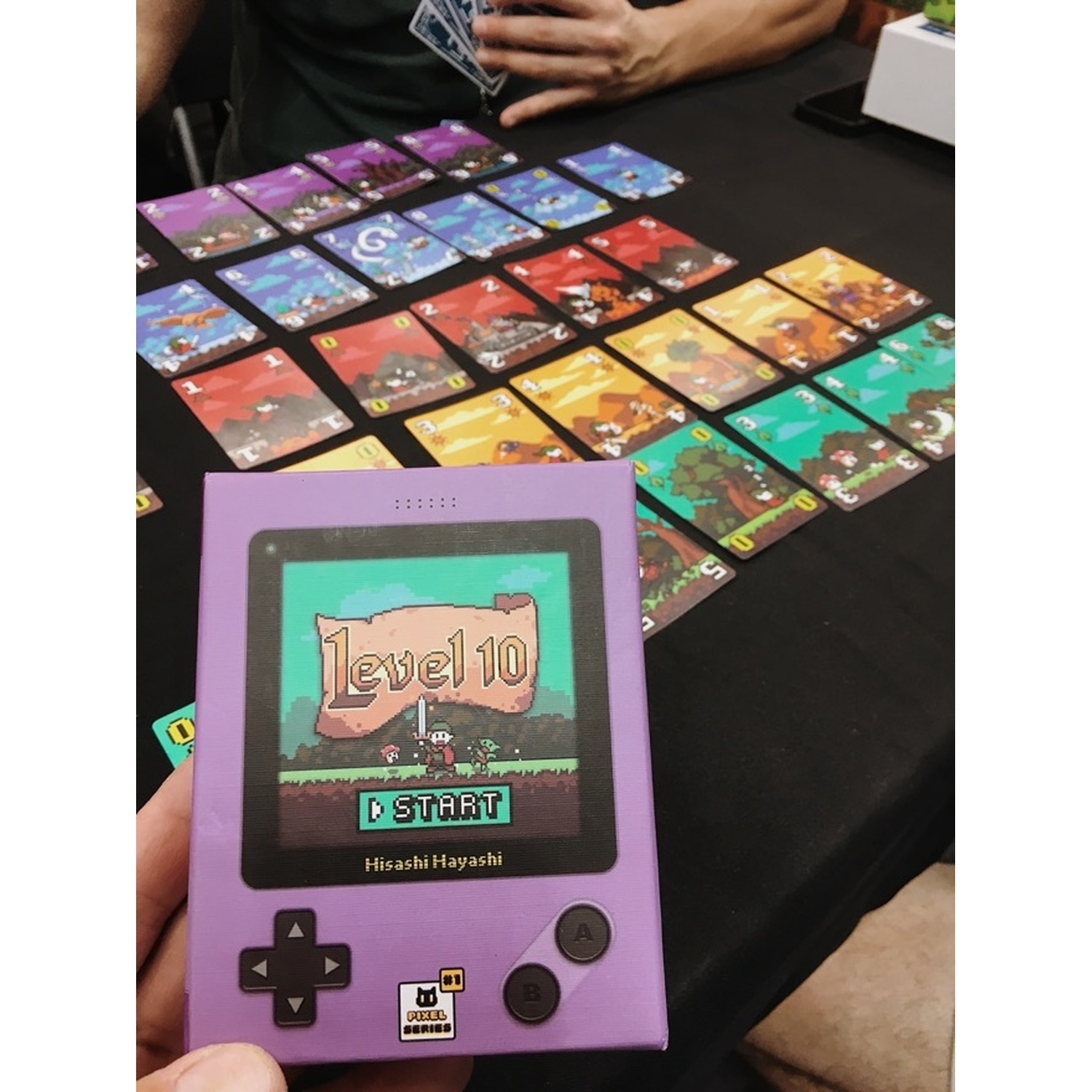 Una console di gioco portatile viola con 'Level 10' e una scena di gioco. Le carte sono disposte su una superficie nera.