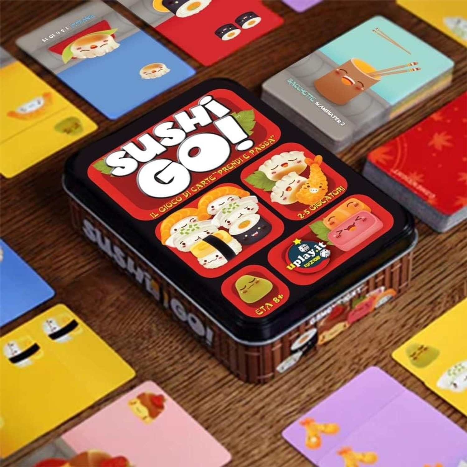 Scatola del gioco di carte Sushi Go! circondata da carte con illustrazioni di sushi. Sfondo tavolo di legno.