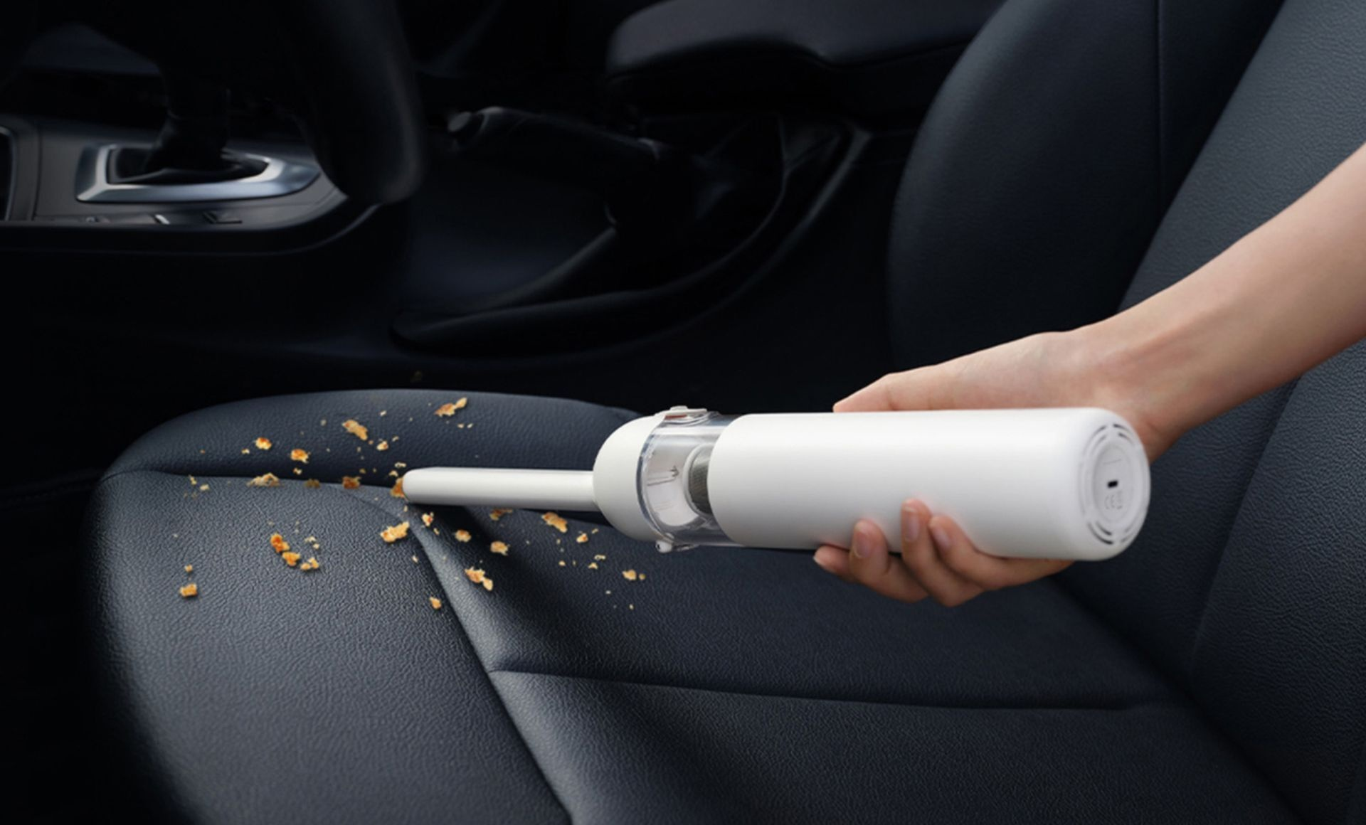 Ręka trzyma biały odkurzacz i czyści okruchy na czarnym siedzeniu samochodowym. W tle wnętrze samochodu.