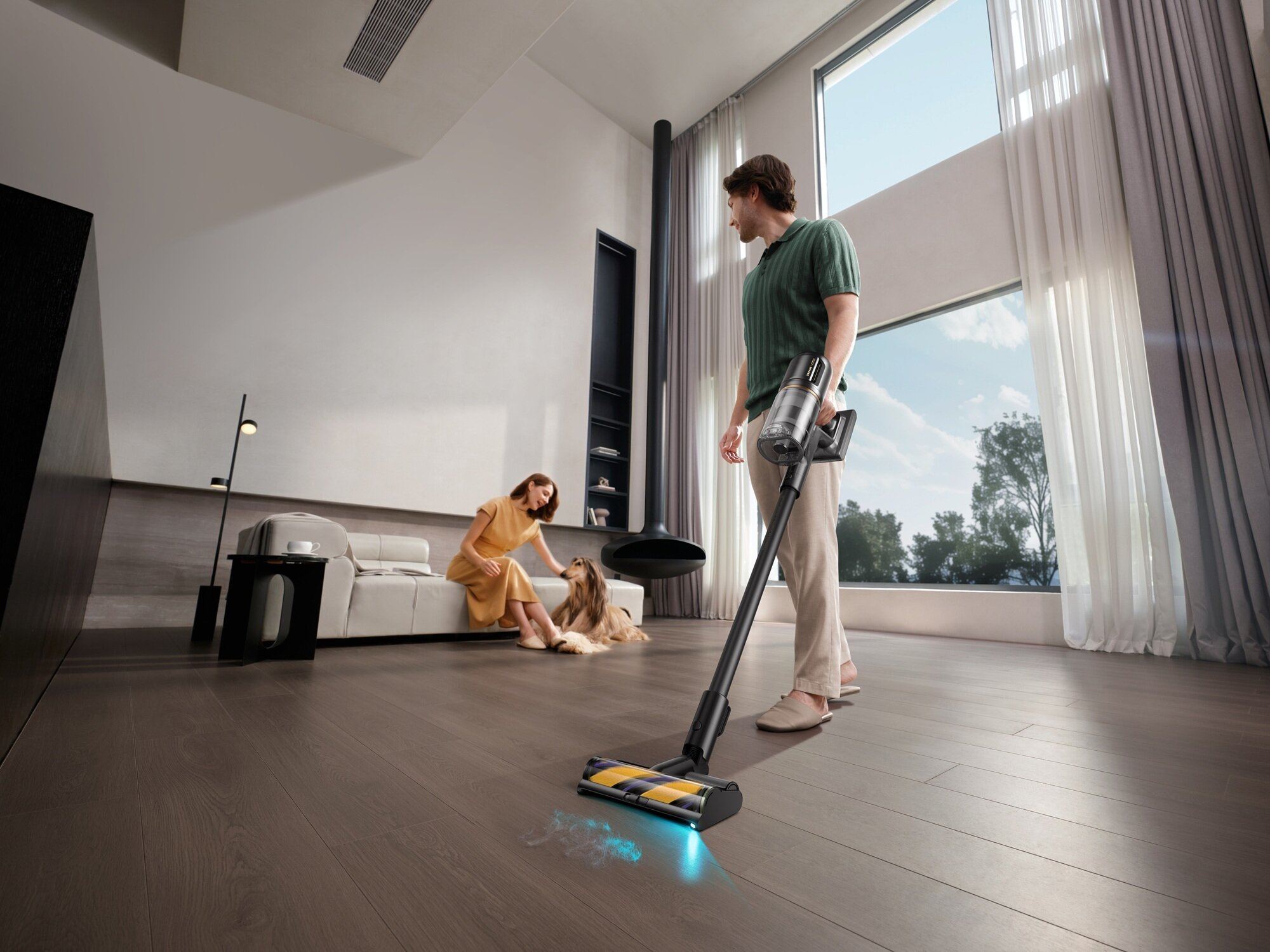 Mężczyzna odkurza drewnianą podłogę. Kobieta siedzi z psem na kanapie. Duże okno z zasłonami w tle.