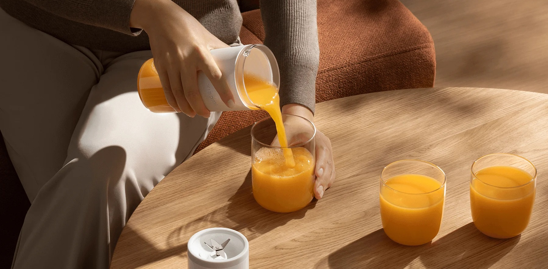 Osoba wlewa sok pomarańczowy z blendera do szklanki. Dwie napełnione szklanki i podstawa blendera znajdują się na drewnianym stole.