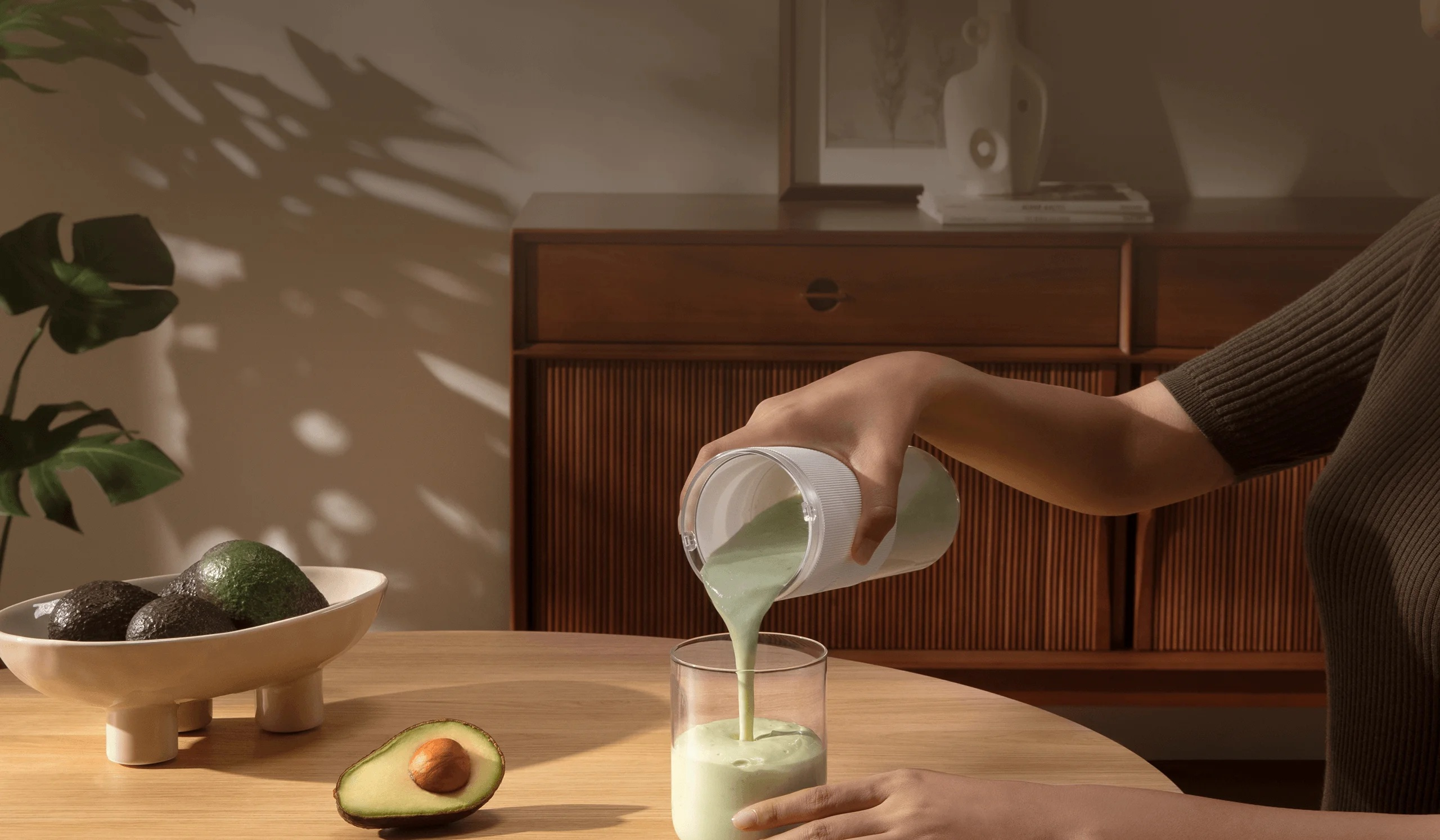 Osoba wlewa zielone smoothie z blendera do szklanki. Awokado leżą na stole. W tle drewniana szafka.