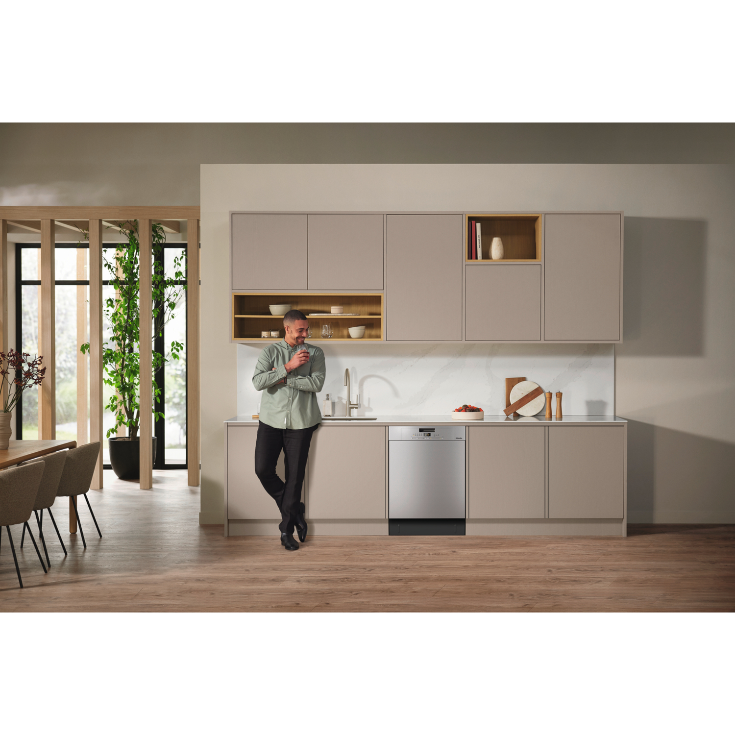 A man leans by a kitchen counter, smiling. A dishwasher is visible. The kitchen has light-colored cabinets and wooden floors.