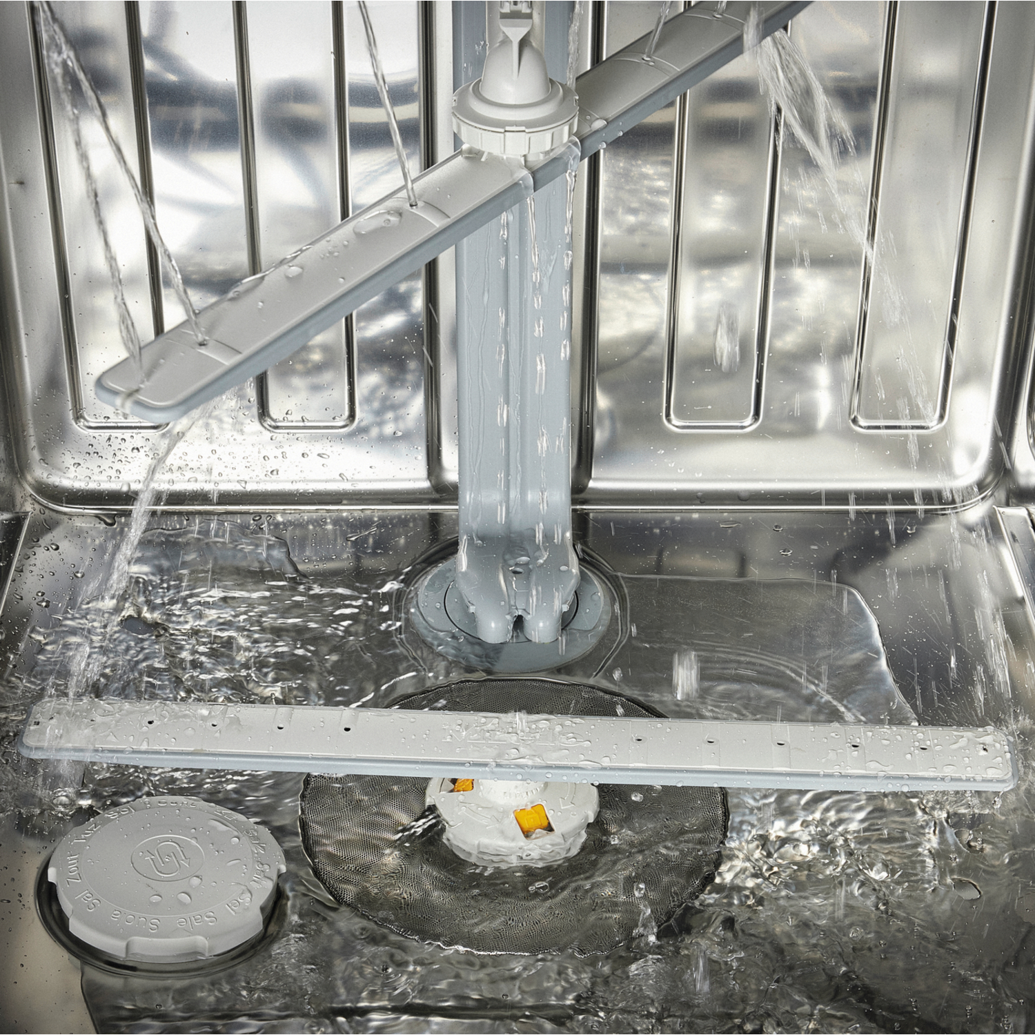 Inside a dishwasher with water spraying. Gray arms spray water. Water is splashing. The inside is stainless steel. A white filter is visible.