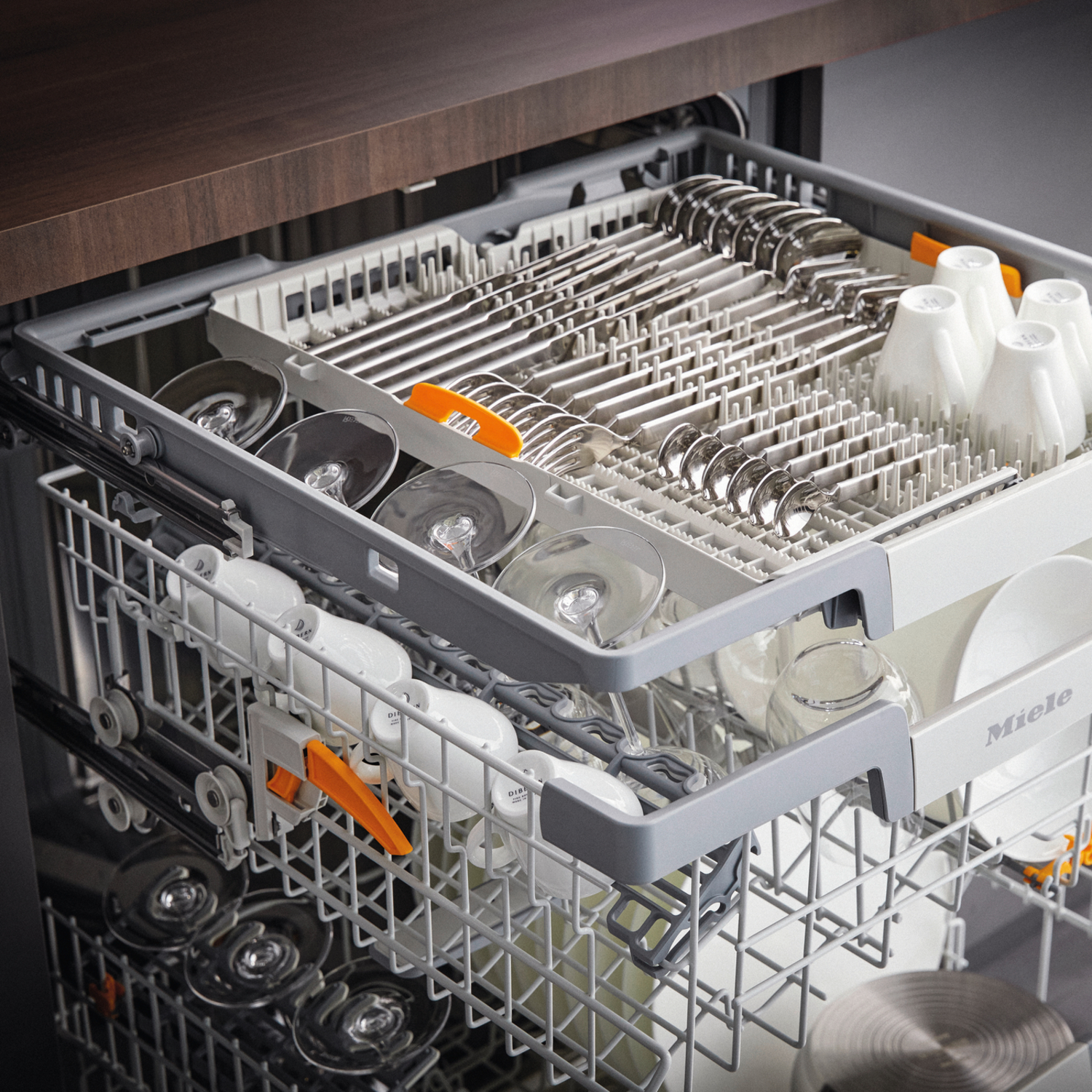 Dishwasher interior loaded with silverware, glasses, and white cups. The top rack holds silverware, and the bottom has glasses.