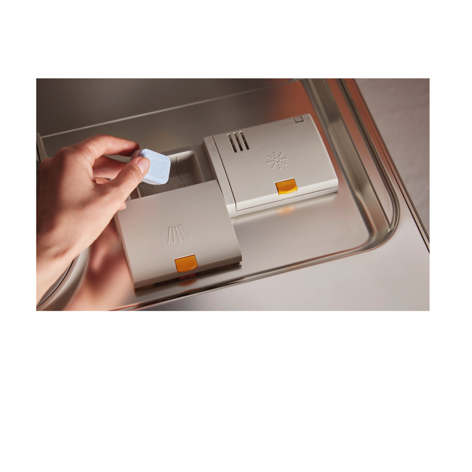 Hand placing a blue dishwasher tablet into the dispenser. Two beige dispenser compartments are visible on a metal surface.