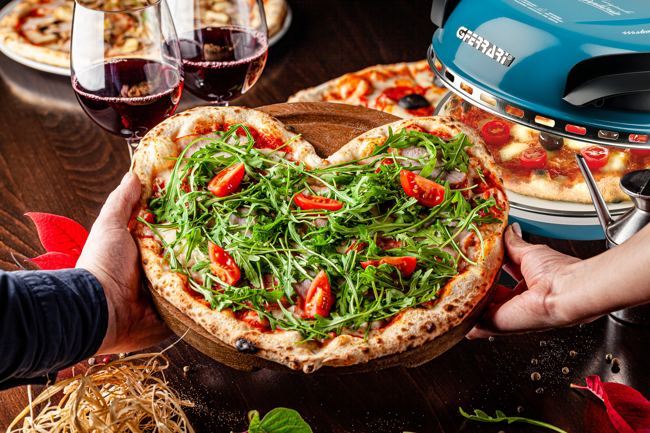Herzförmige Pizza mit Rucola und Tomaten wird gehalten. Andere Pizzen, Rotwein und ein blauer Pizzaofen sind ebenfalls zu sehen.