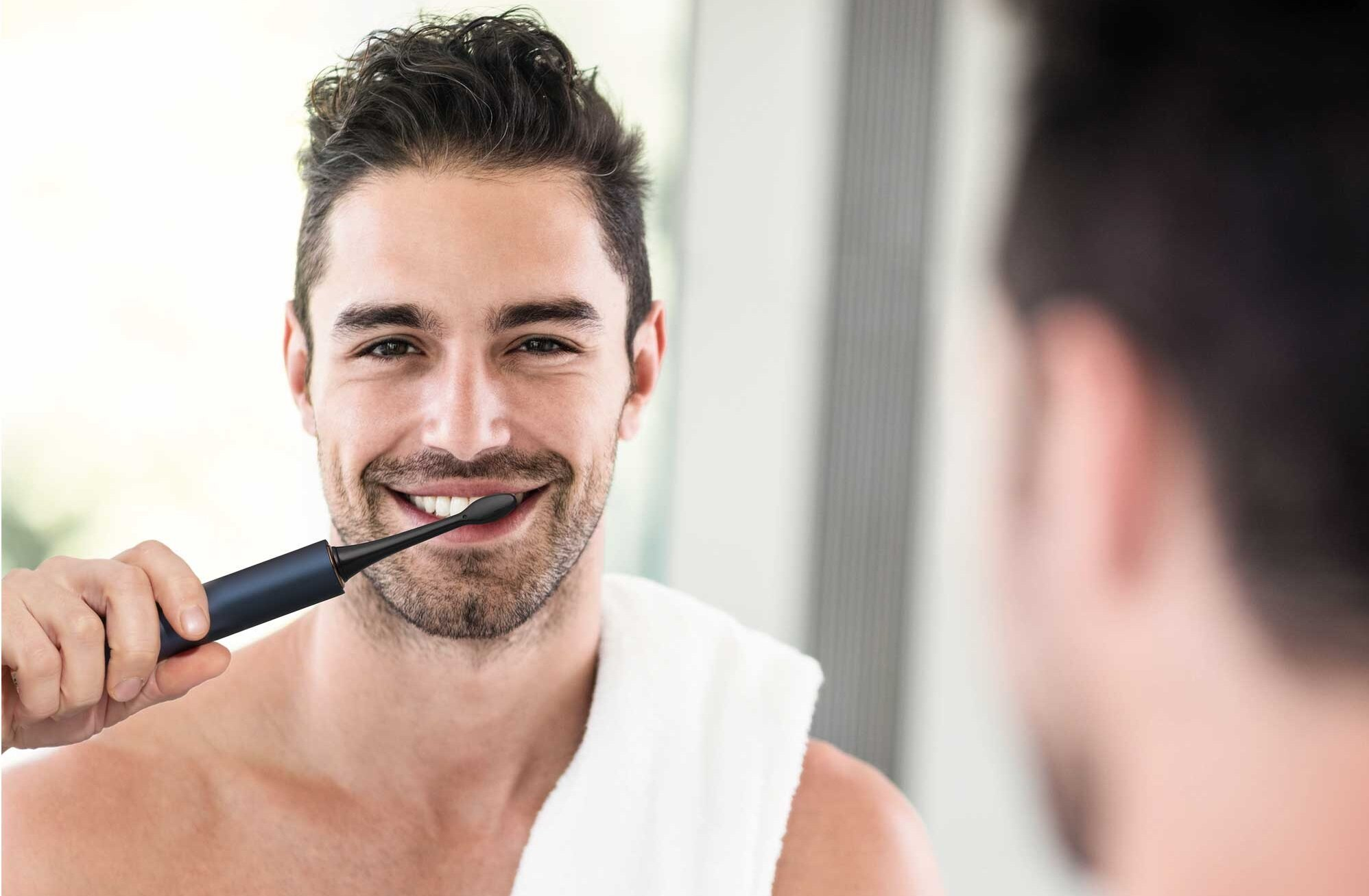 Mężczyzna myje zęby niebieską szczoteczką elektryczną, uśmiechając się do lustra. Biały ręcznik wisi na jego ramieniu.