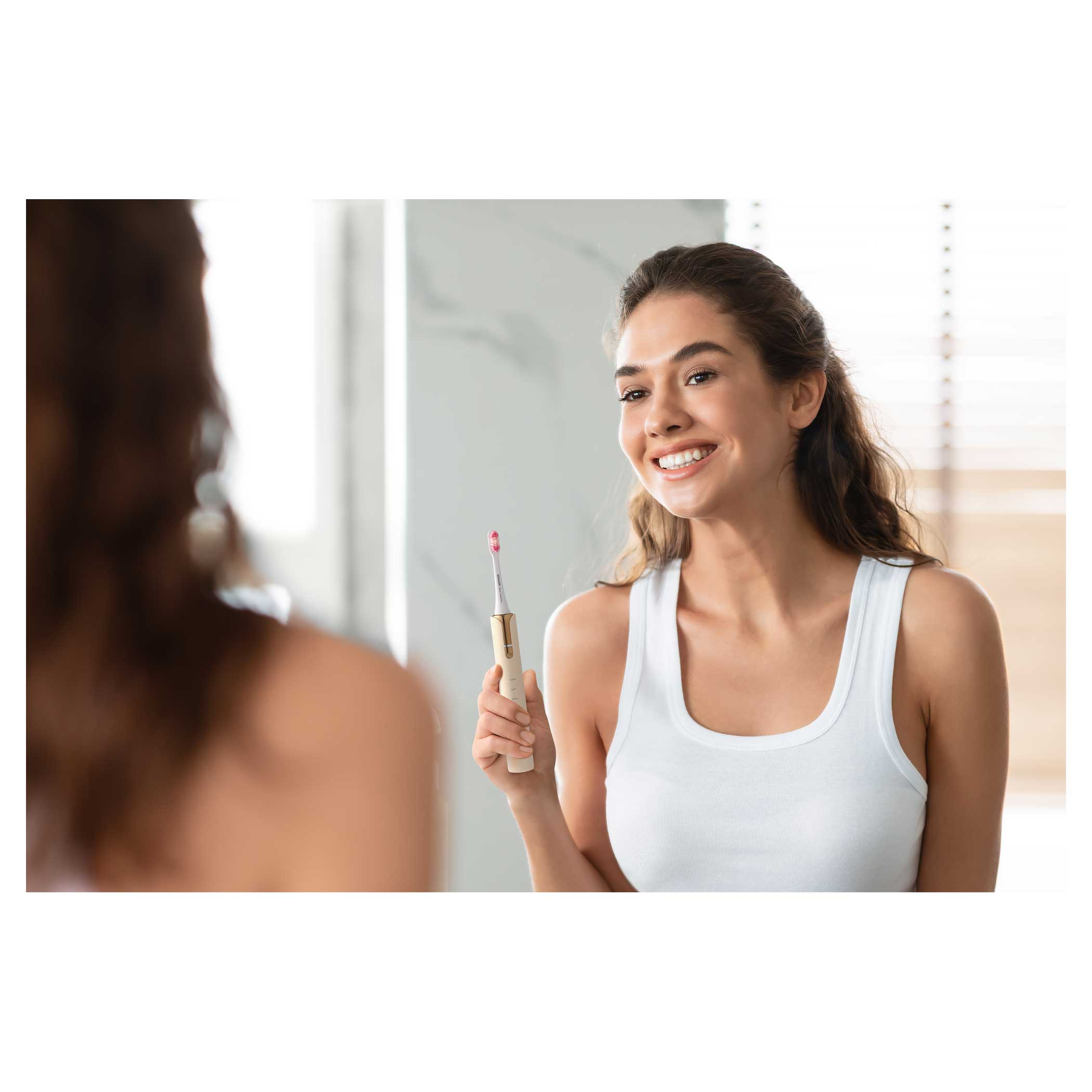 Kobieta uśmiecha się w lustrze, trzyma szczoteczkę. Tło: okno i rozmyte odbicie.