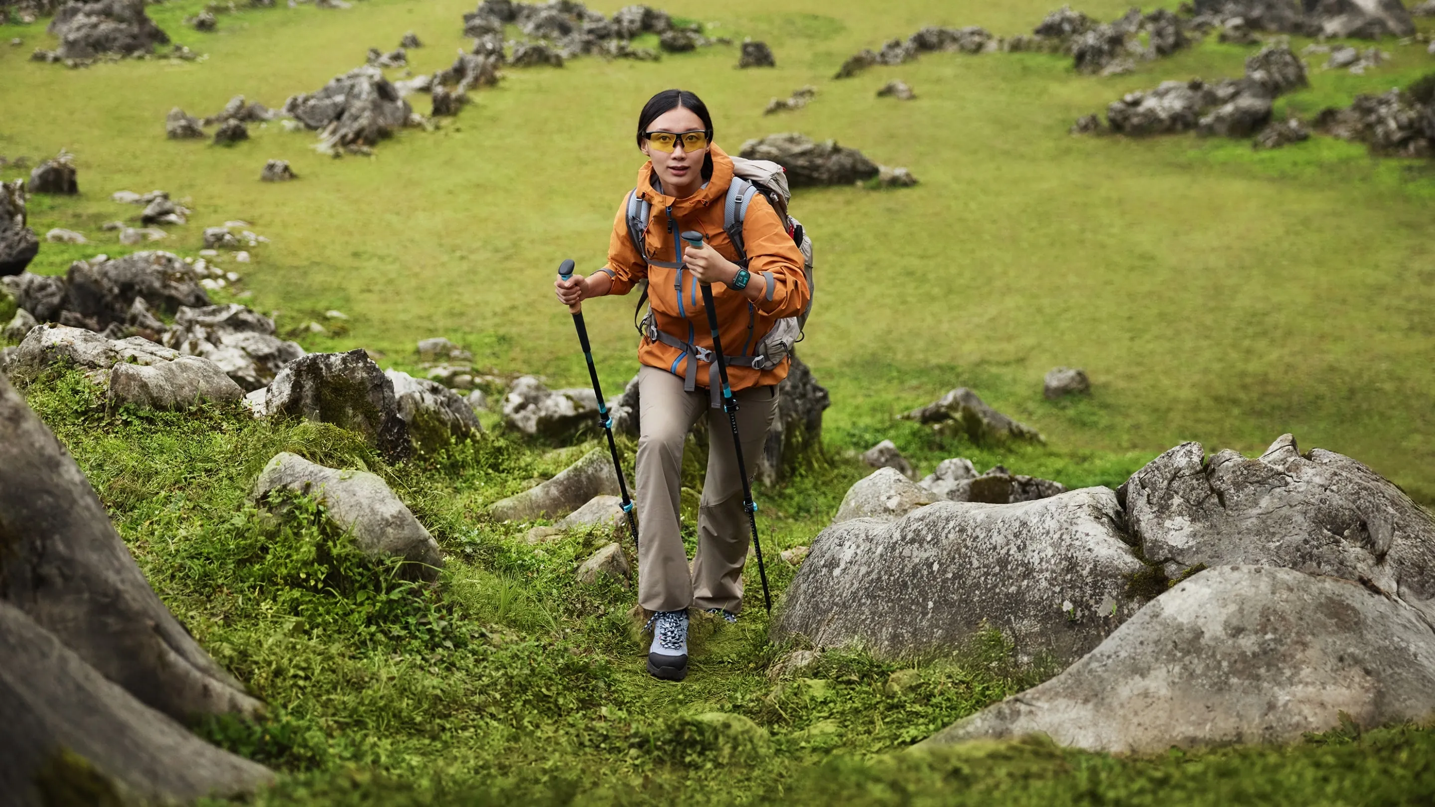 Eine Frau wandert mit Wanderstöcken einen grünen, grasbewachsenen Hang hinauf. Felsen befinden sich im Vorder- und Hintergrund.