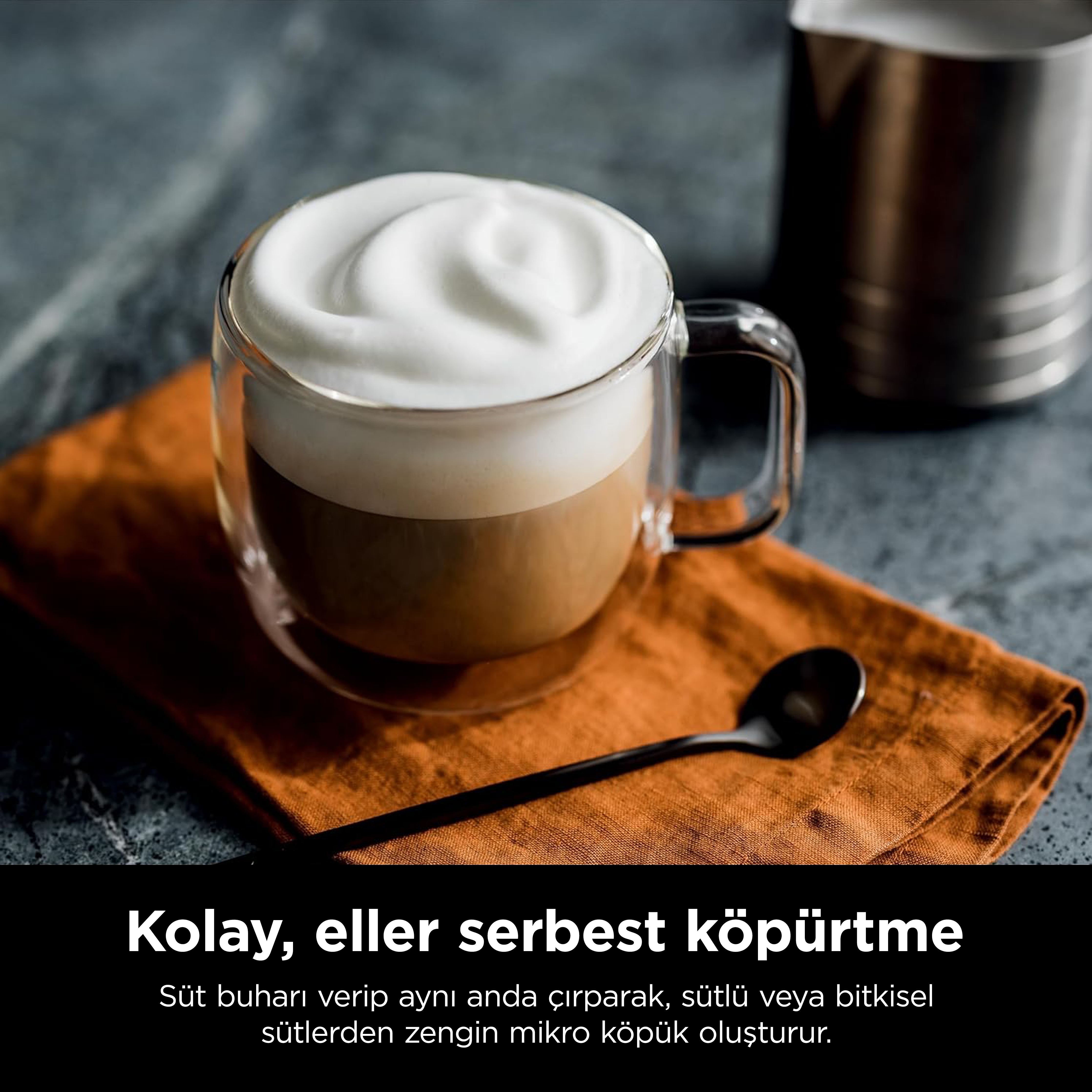 Turuncu bir bez üzerinde kalın köpüklü bir bardak latte, siyah bir kaşık ve çelik bir sütlük.