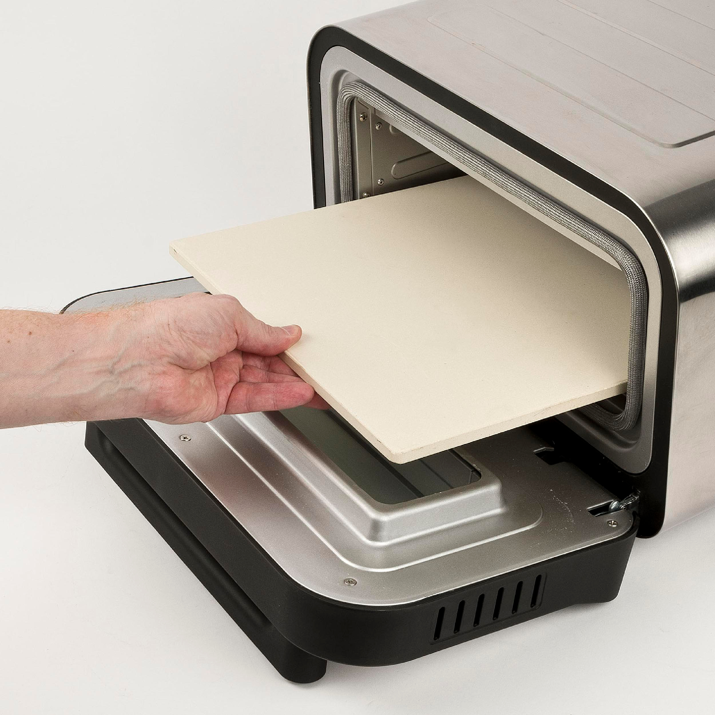 Eine Hand schiebt einen hellfarbigen Pizzastein in einen silber-schwarzen Toaster-Ofen vor weißem Hintergrund.
