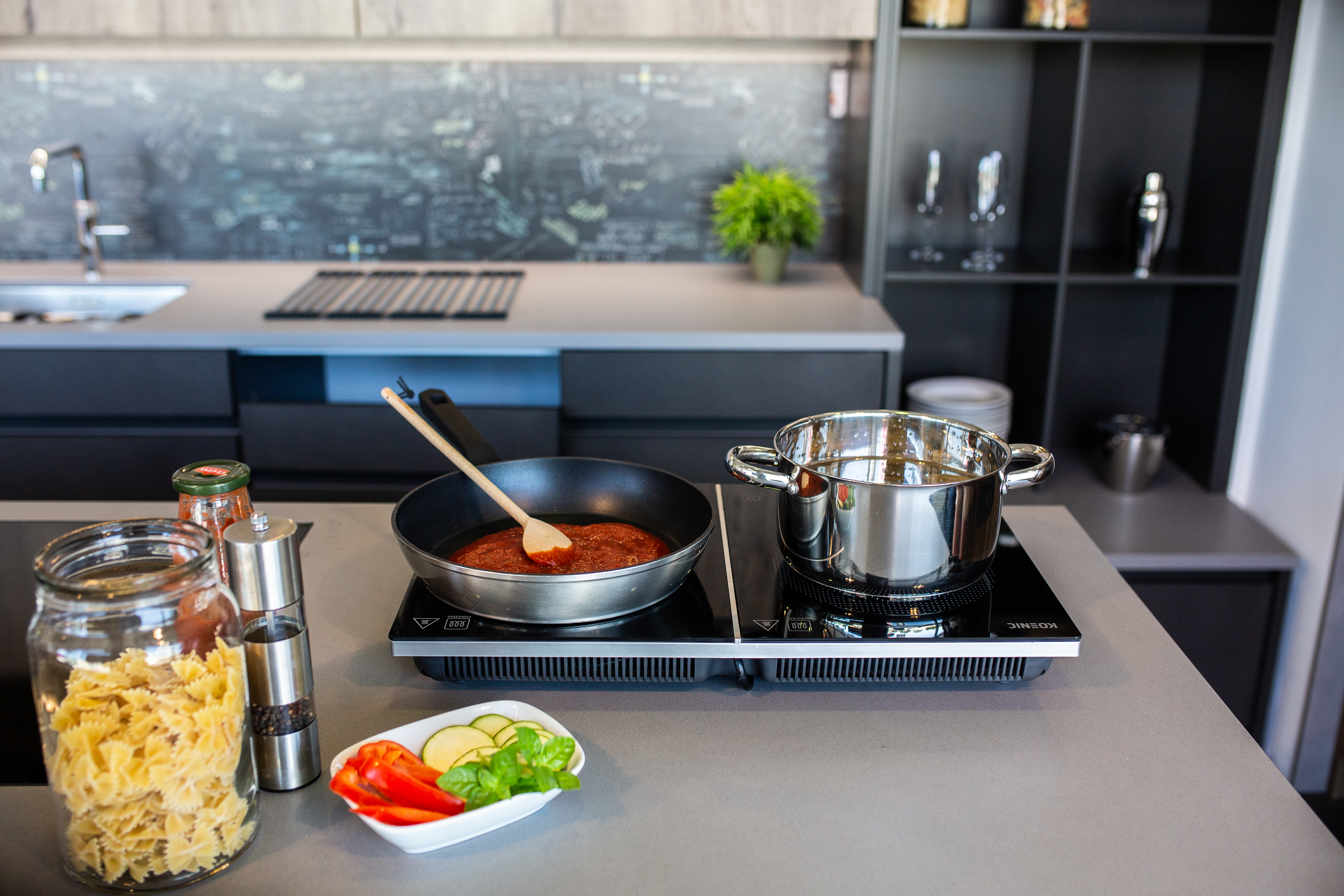 Scena kuchenna z przyborami do gotowania, makaronem, warzywami i garnkiem z sosem na kuchence elektrycznej. Drewniana łyżka miesza.