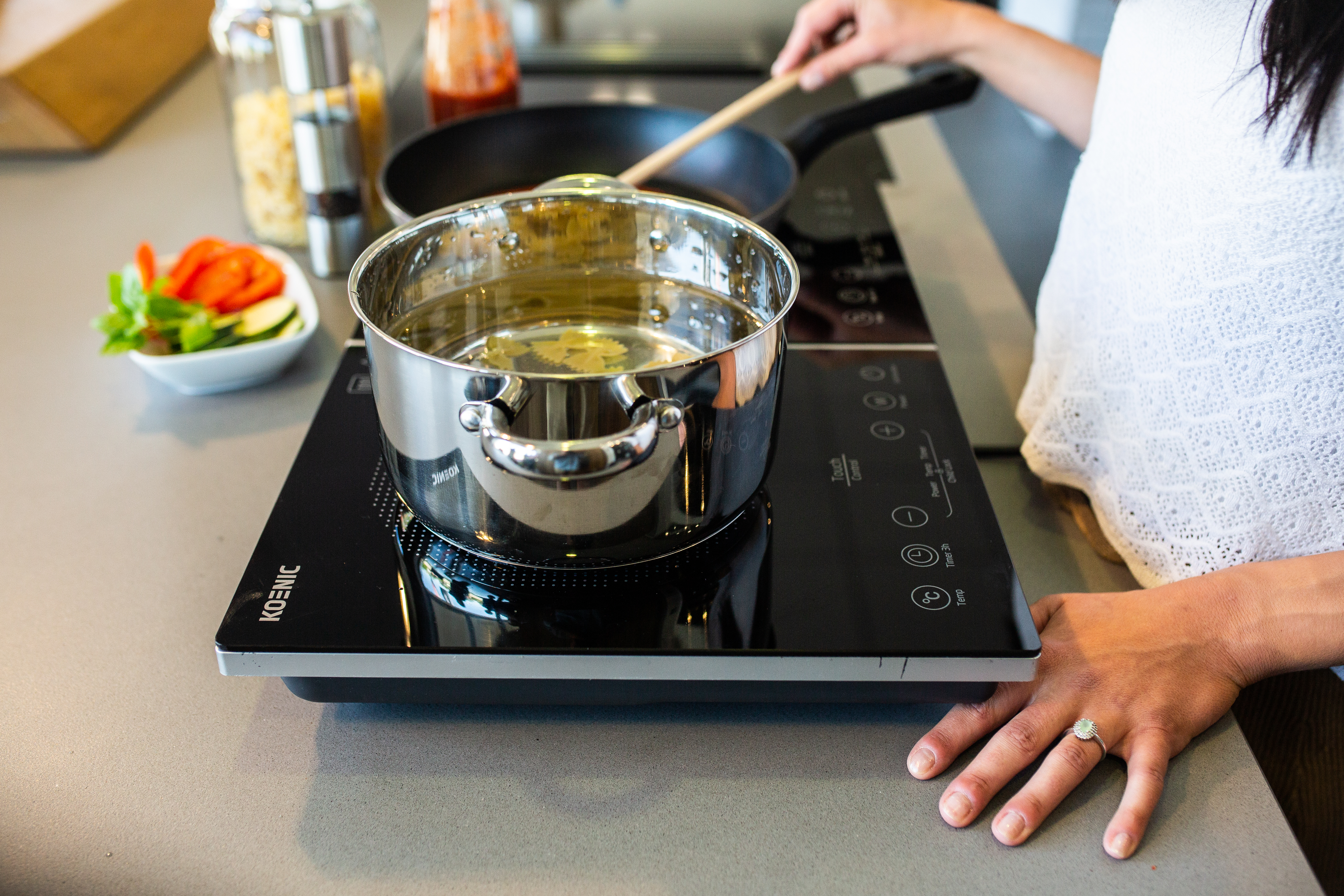 Kobieta gotuje na czarnej płycie indukcyjnej. Widoczny garnek z wodą, patelnia i warzywa. Kobieta trzyma drewnianą łyżkę.