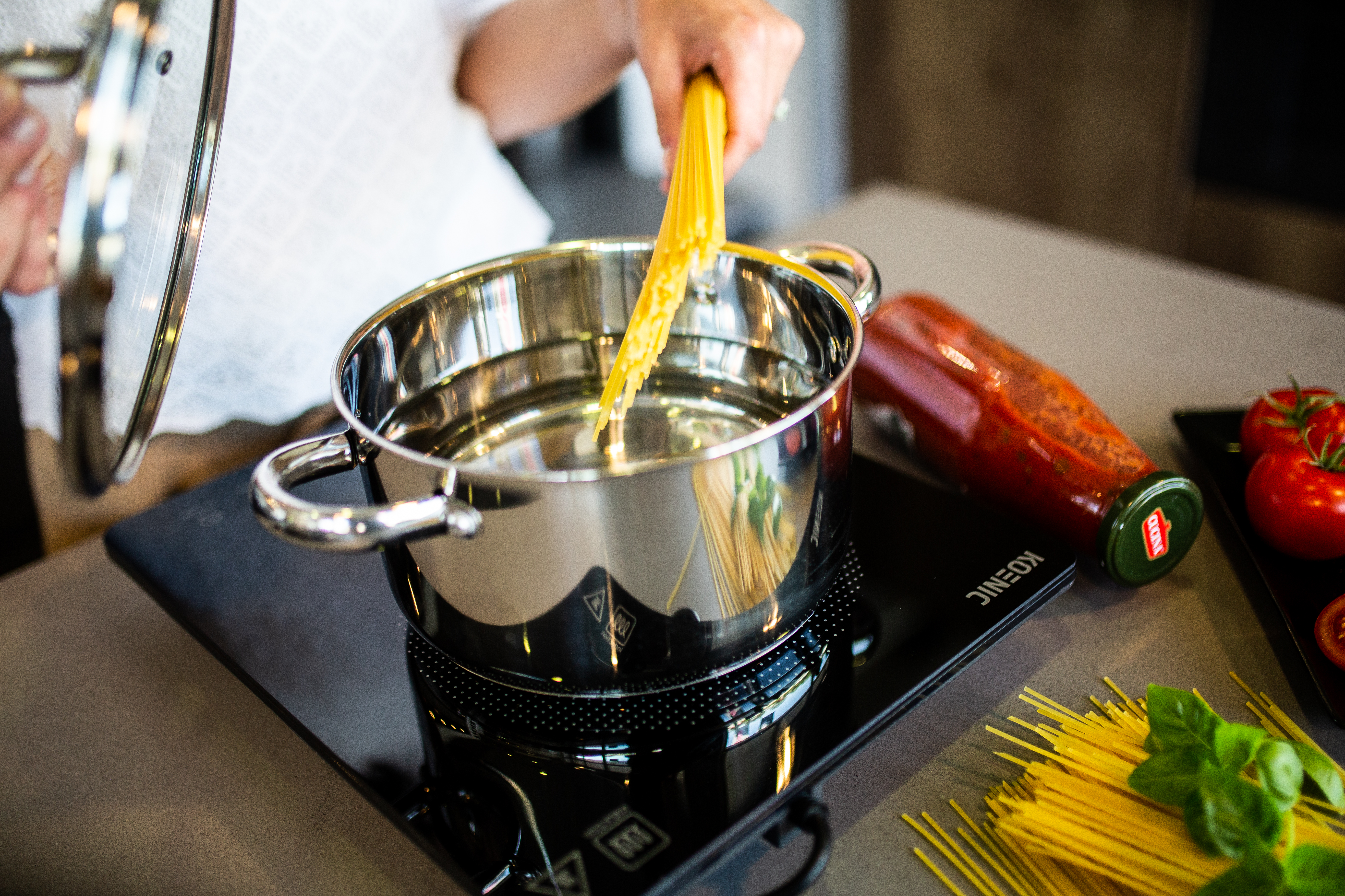 Ręka dodaje spaghetti do wrzącej wody w garnku. Pomidory, sos i makaron na blacie.
