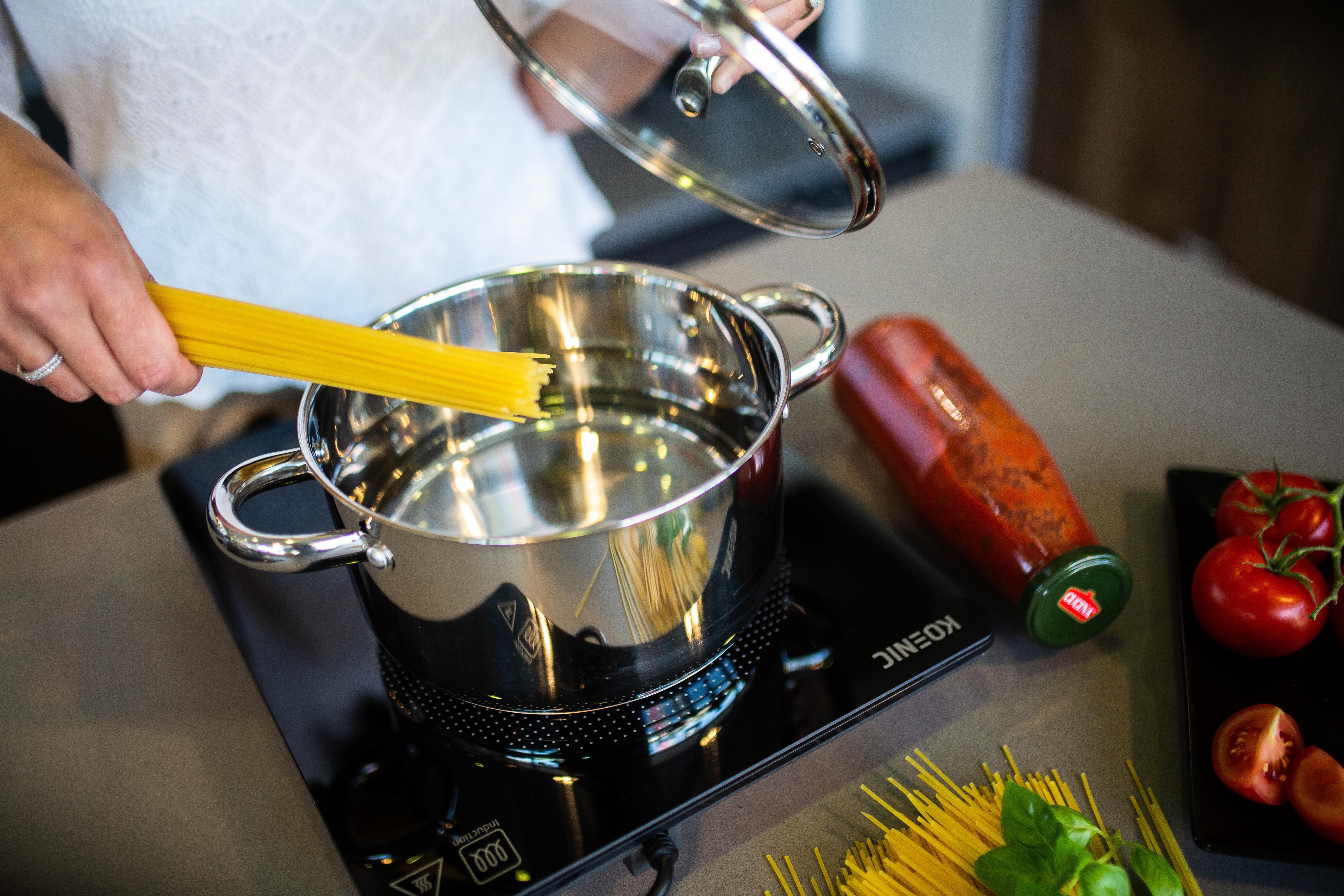 Osoba dodaje spaghetti do garnka z wrzącą wodą. Składniki to pomidory, sos i bazylia. Trzymana jest szklana pokrywka.