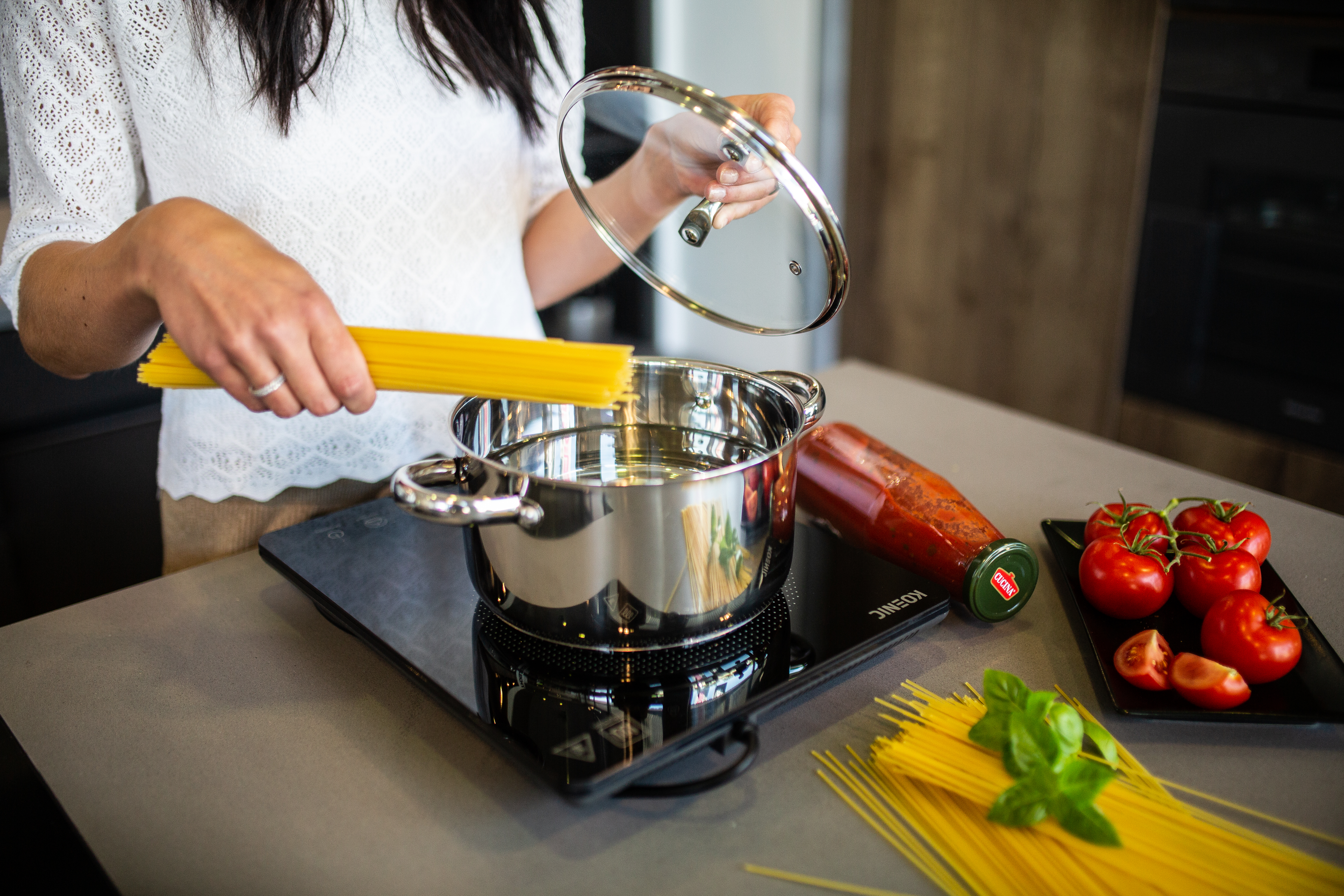 Osoba dodaje makaron do garnka na kuchence. W pobliżu są pomidory, sos i bazylia.