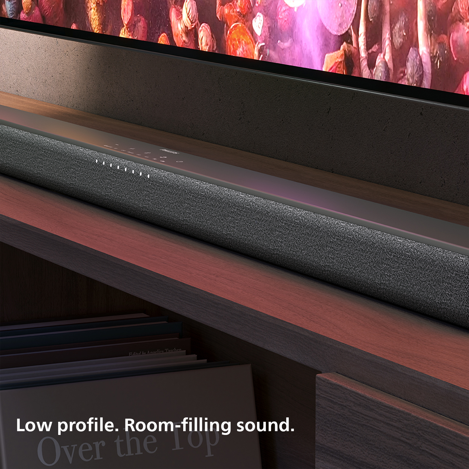 A soundbar on a wooden shelf, below a TV. Books are on the shelf below the soundbar. Text reads: Low profile. Room-filling sound.