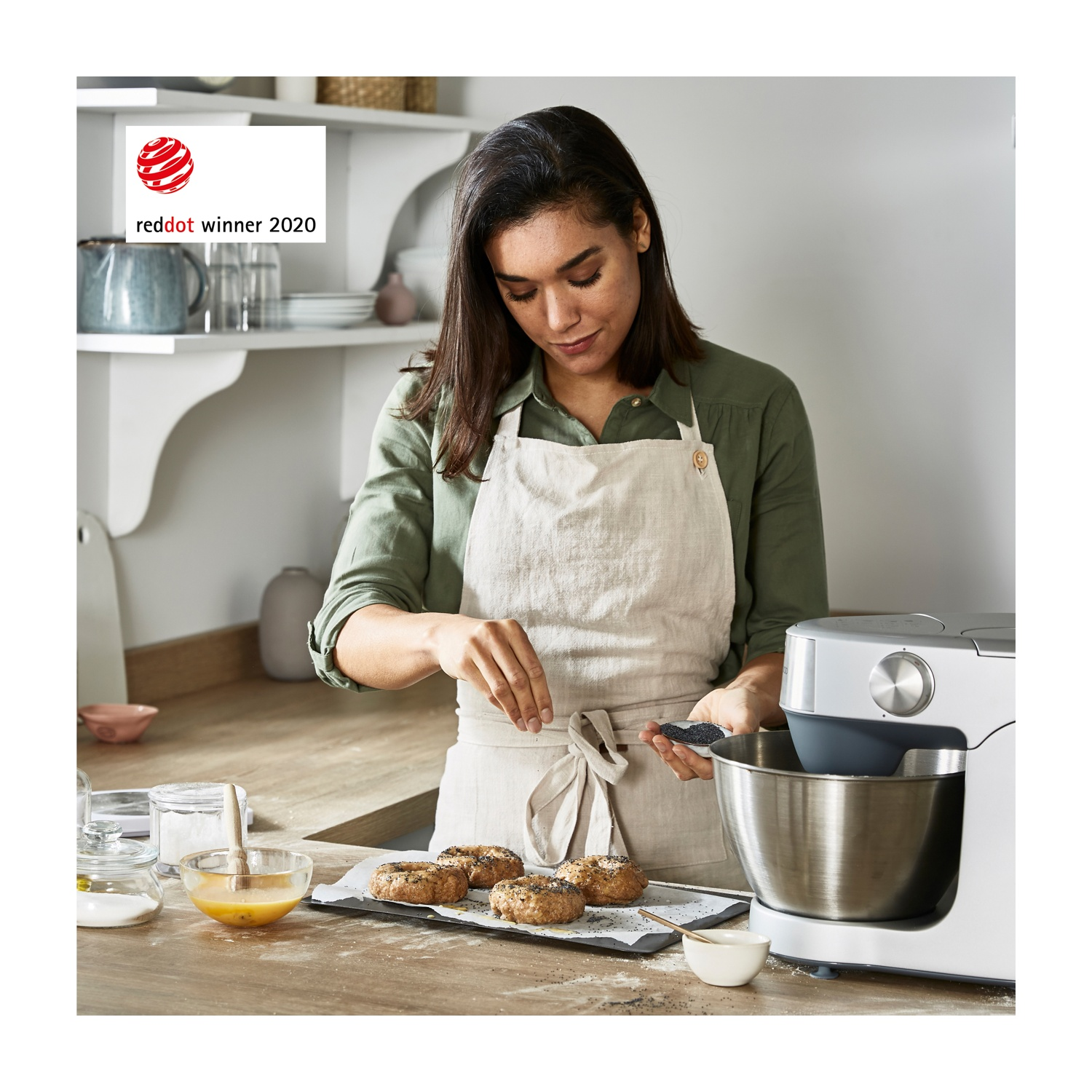 Una donna con un grembiule cosparge semi sui bagel. Un robot da cucina a destra. Logo Reddot winner 2020 in alto a sinistra.